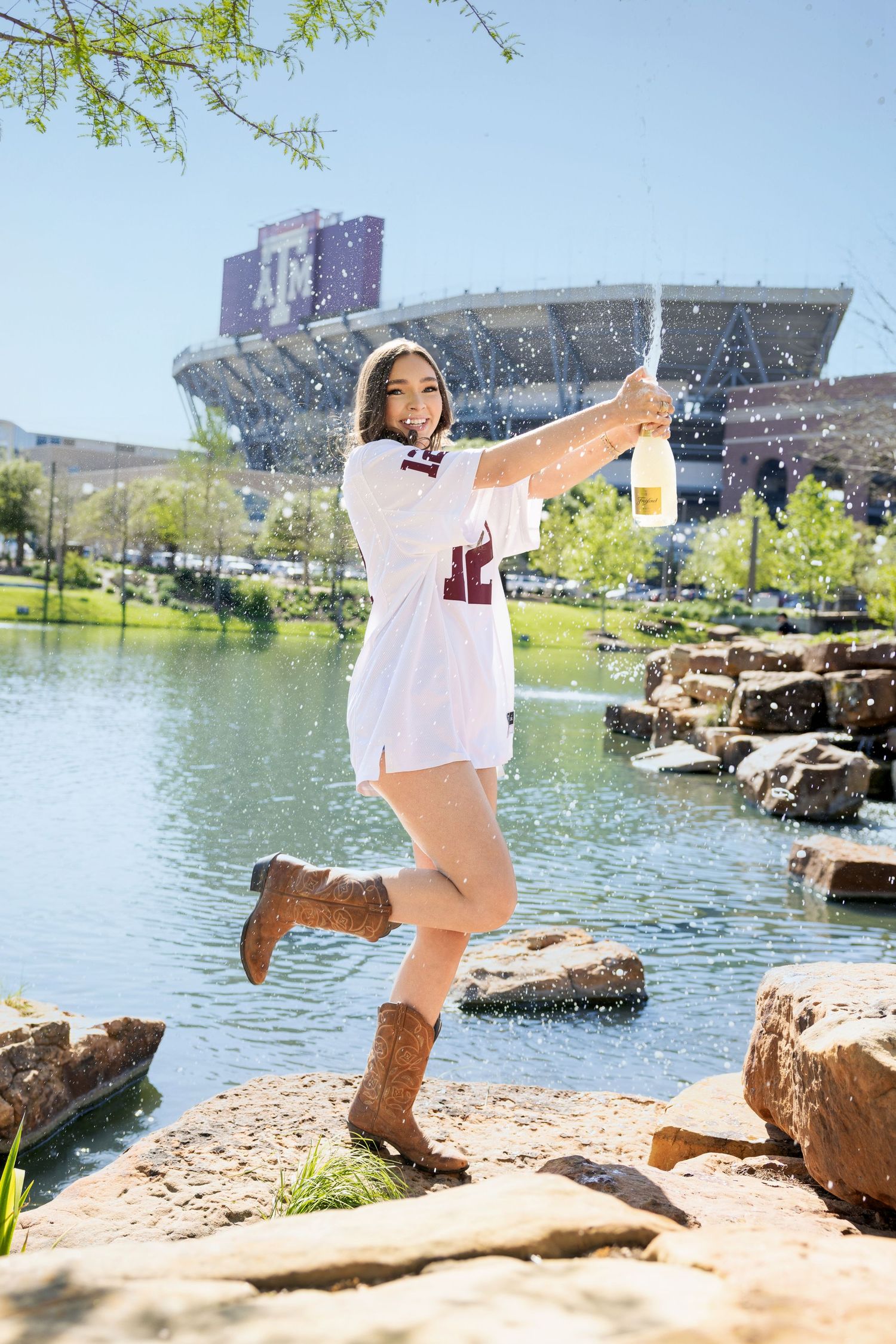 Person wearing white jersey and brown boots poses on rocks by a lake with stadium and trees in background on sunny day.