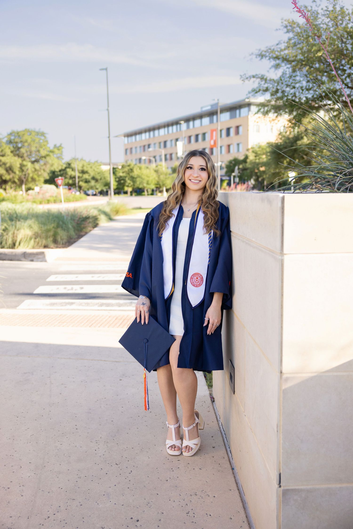 Graduate in blue cap and gown poses with diploma holder against concrete wall on sunny campus walkway.