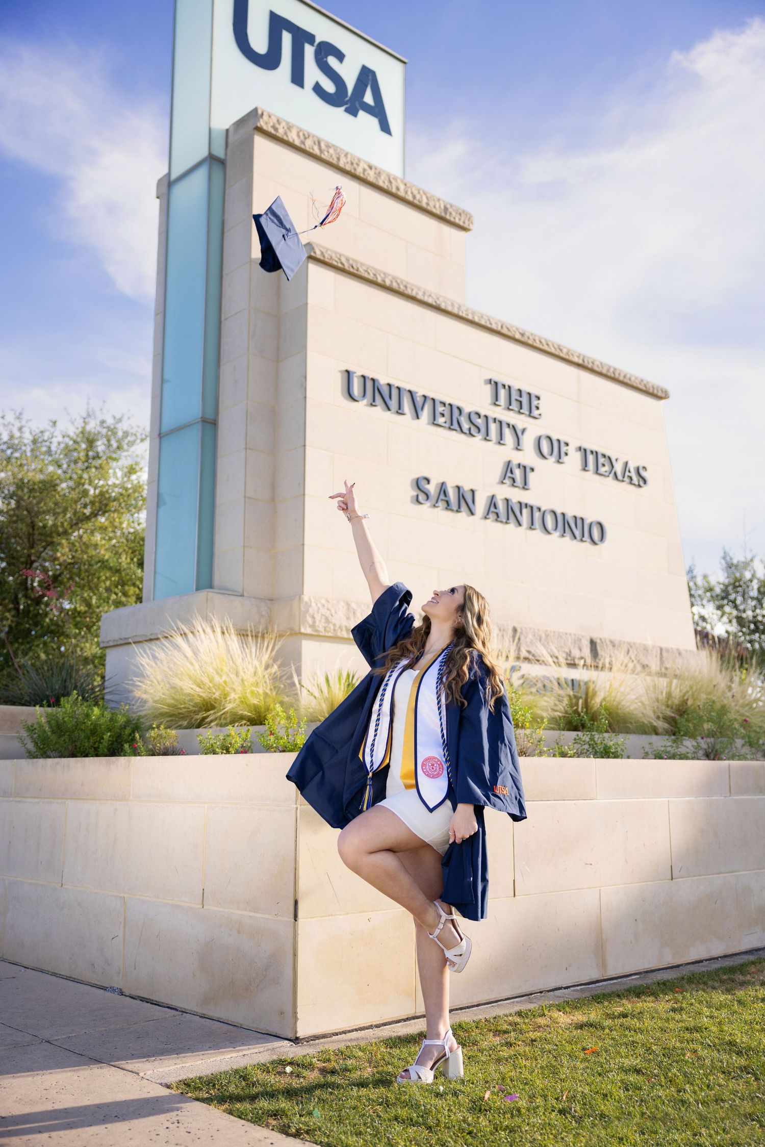 A graduation photo taken at the UTSA entrance sign showing a celebratory pose in academic regalia on a sunny day.
