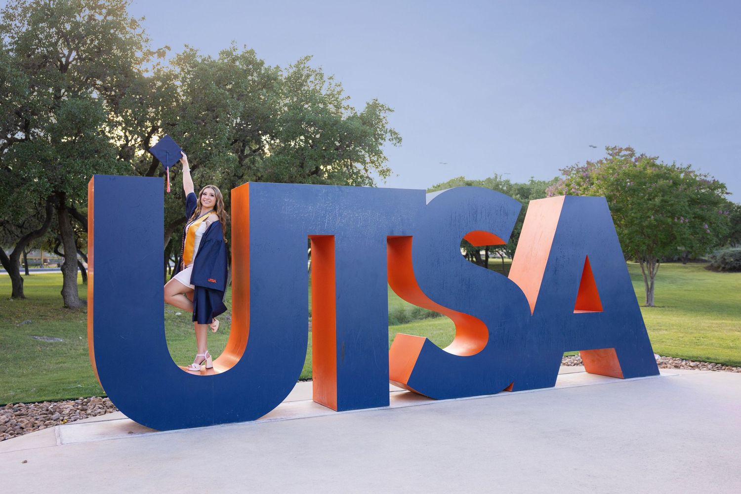 Large blue UTSA block letters installed outdoors on grass with trees in background under blue sky.