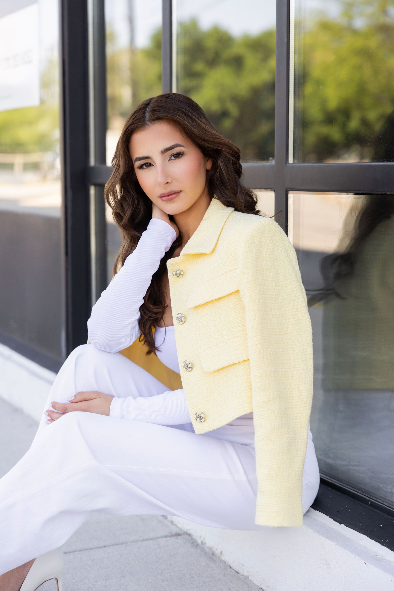 Someone wearing a cropped yellow blazer and white outfit poses elegantly by a window.