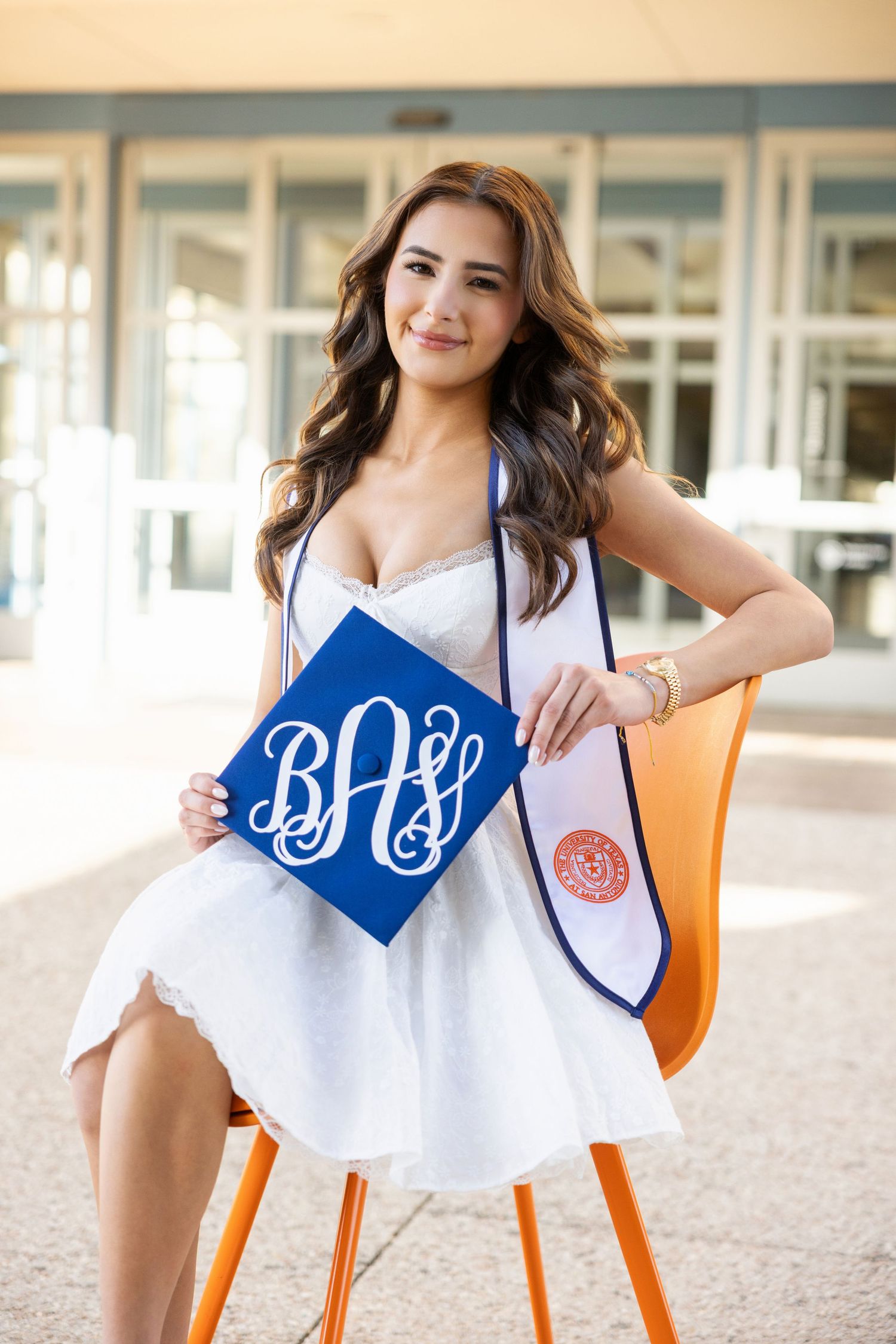 Graduate in white dress sits in orange chair holding blue monogrammed graduation sash and school stole.
