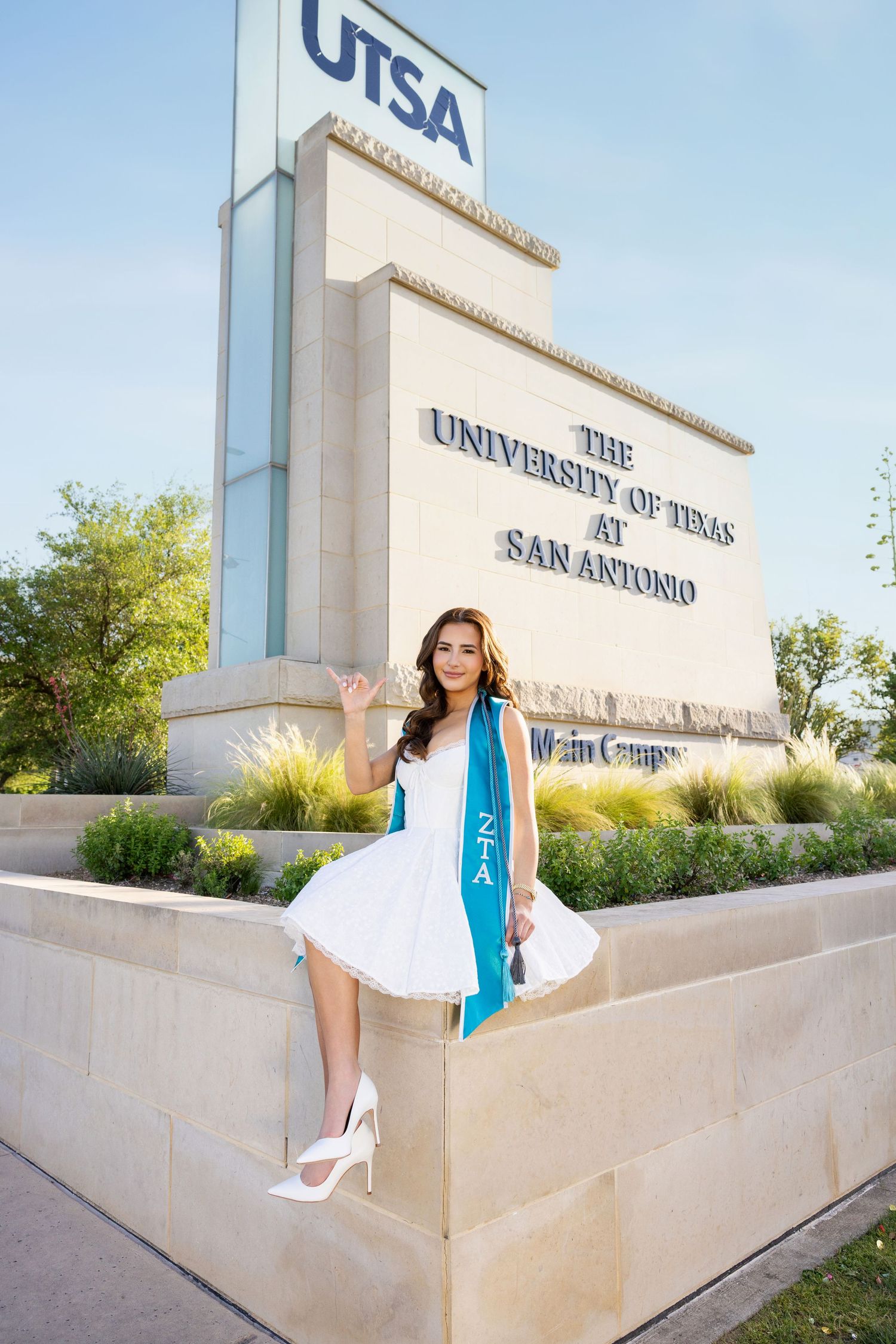 Graduation photo at UTSA campus entrance monument featuring a graduate in white dress and blue sash posing on stone wall.