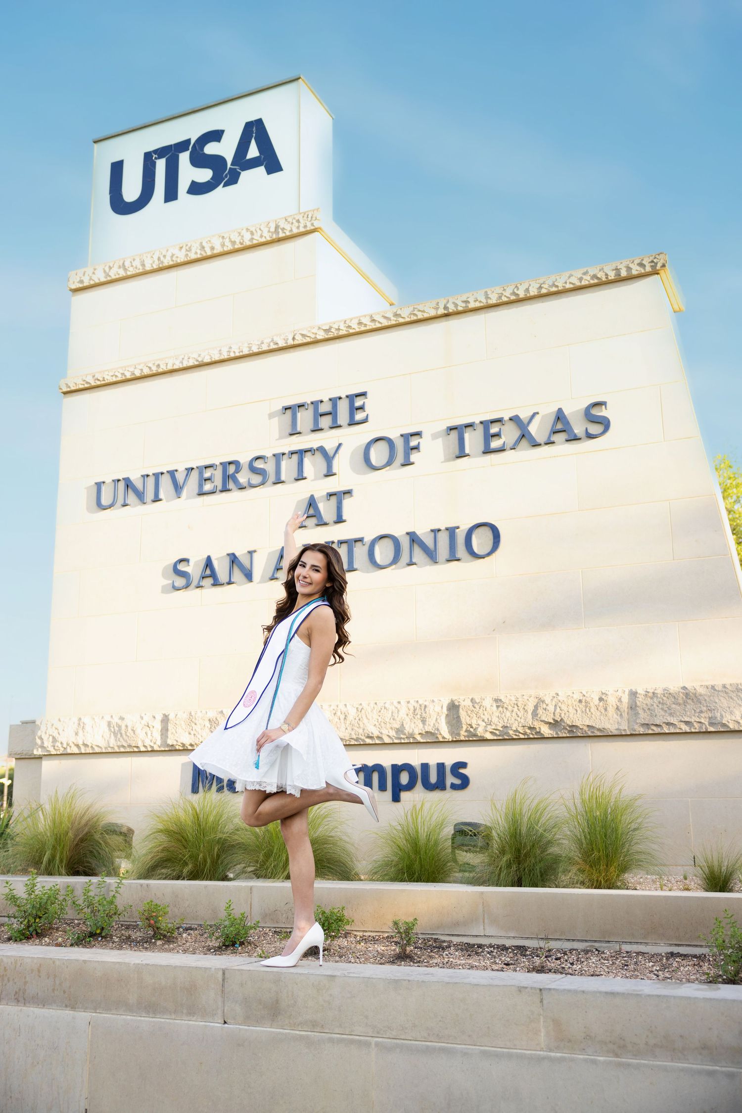 A graduate poses in a white dress by the UTSA (University of Texas at San Antonio) campus entrance sign on a sunny day.