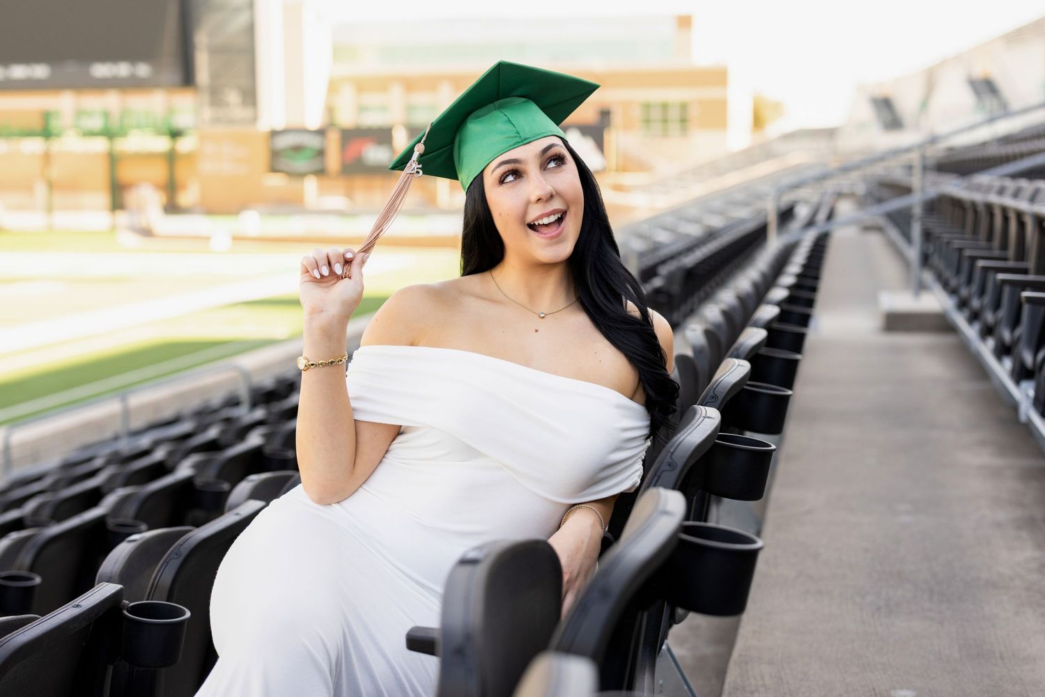 Graduate in white dress and green cap smiling while sitting in stadium bleachers.