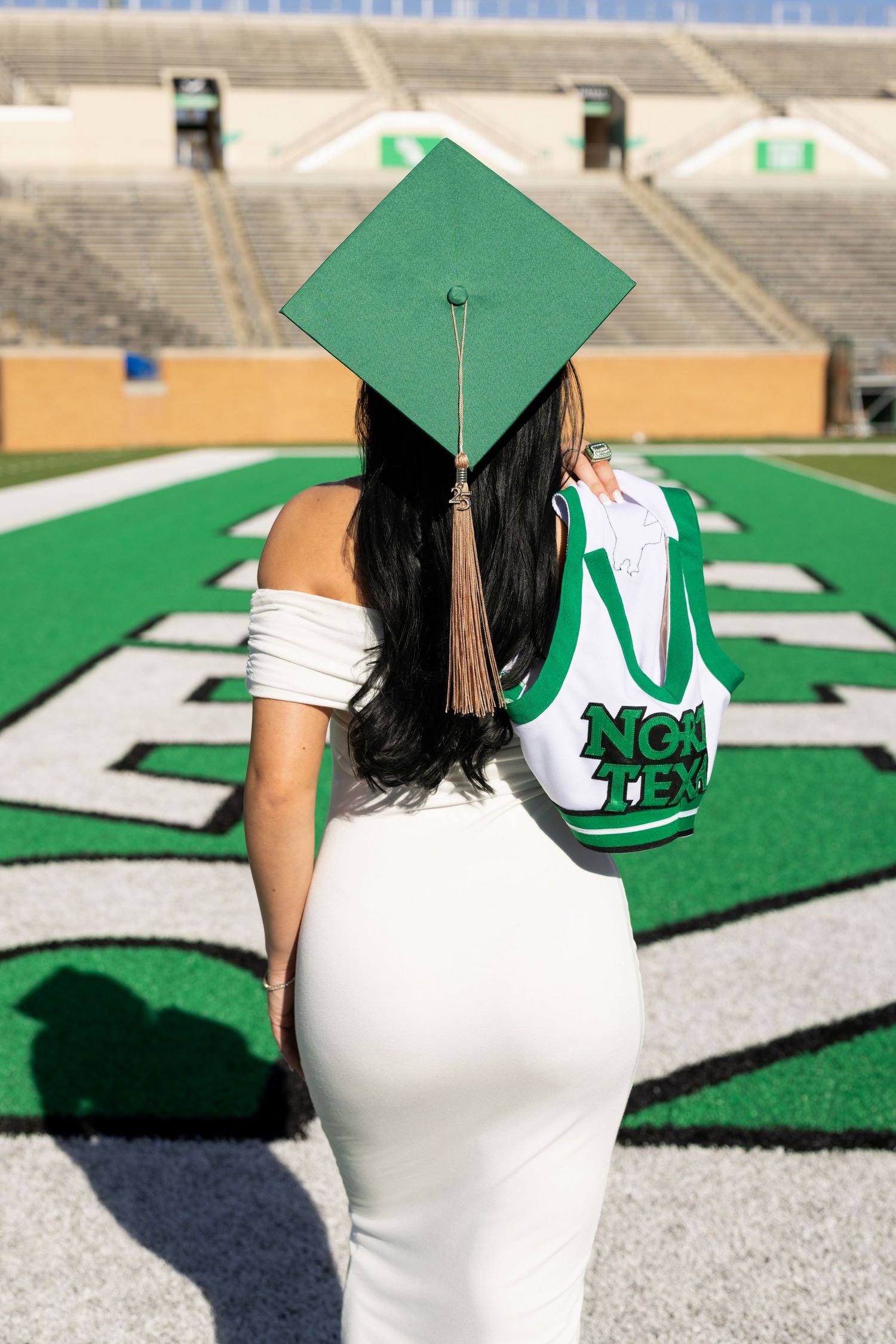 Graduate in white dress and green cap holding spirit gear stands on football field logo in outdoor stadium.