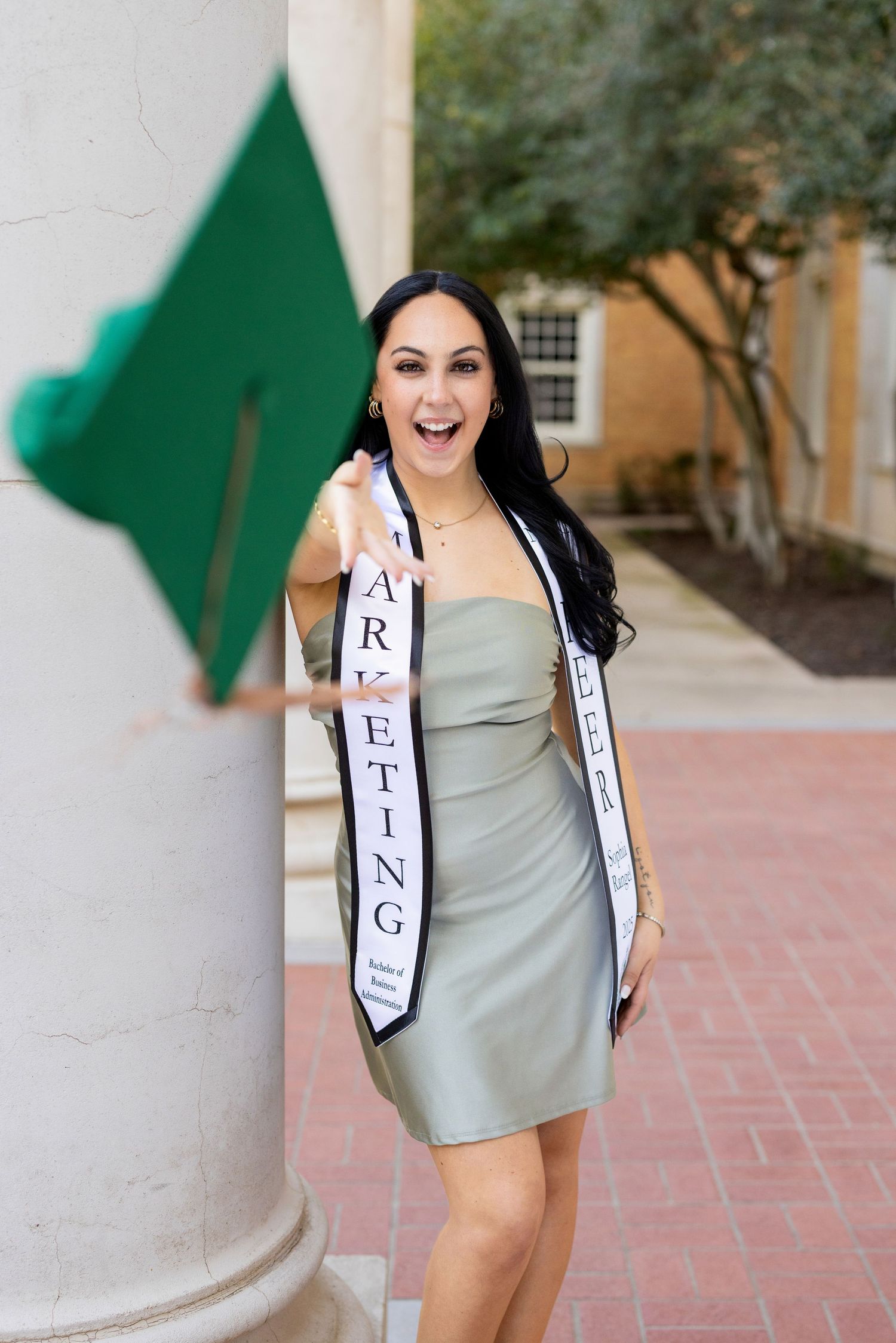 Graduate wearing grey dress and white graduation sash poses excitedly outdoors by column and green banner.