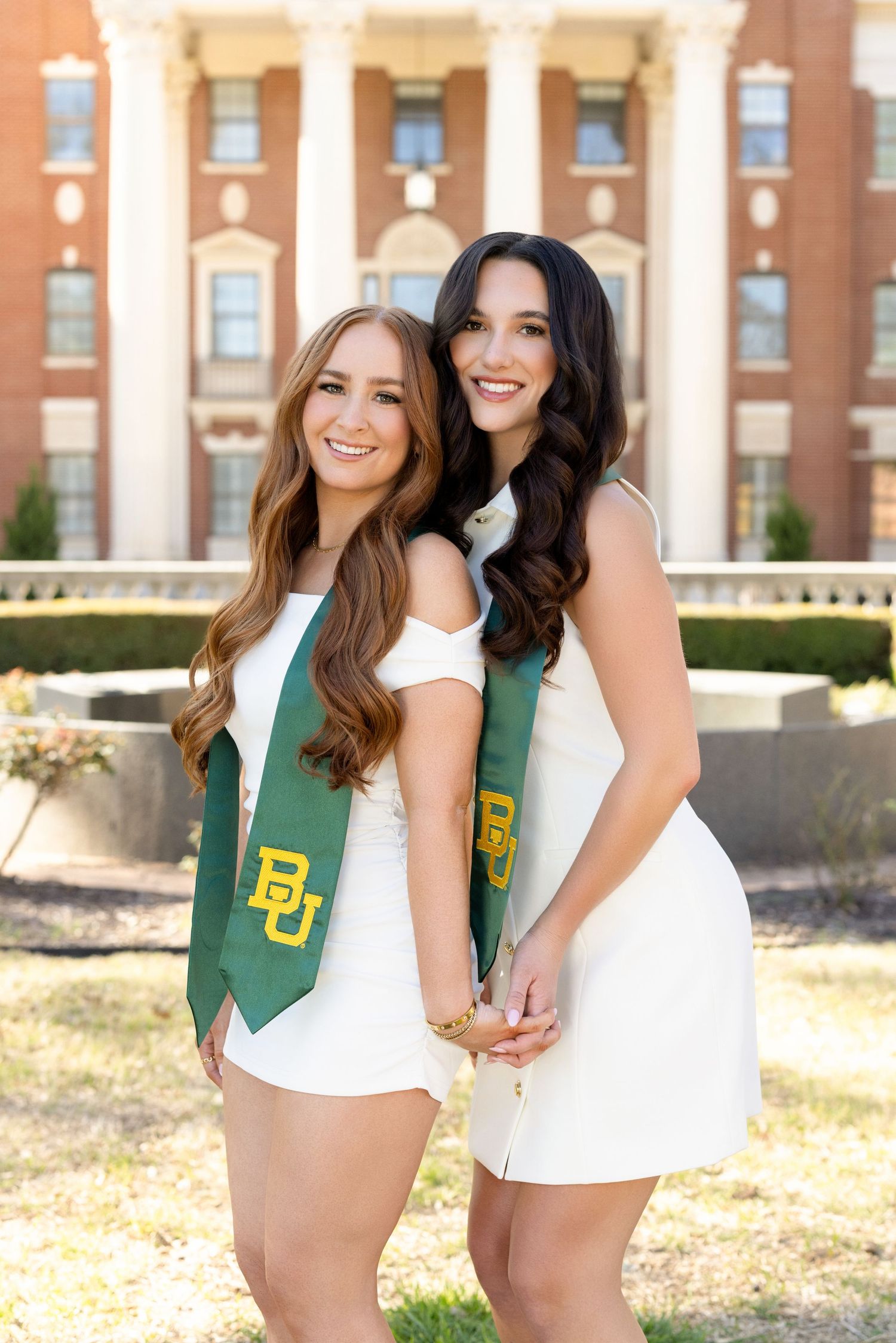 Two students wearing white dresses and green Baylor University graduation stoles pose together on campus grounds.