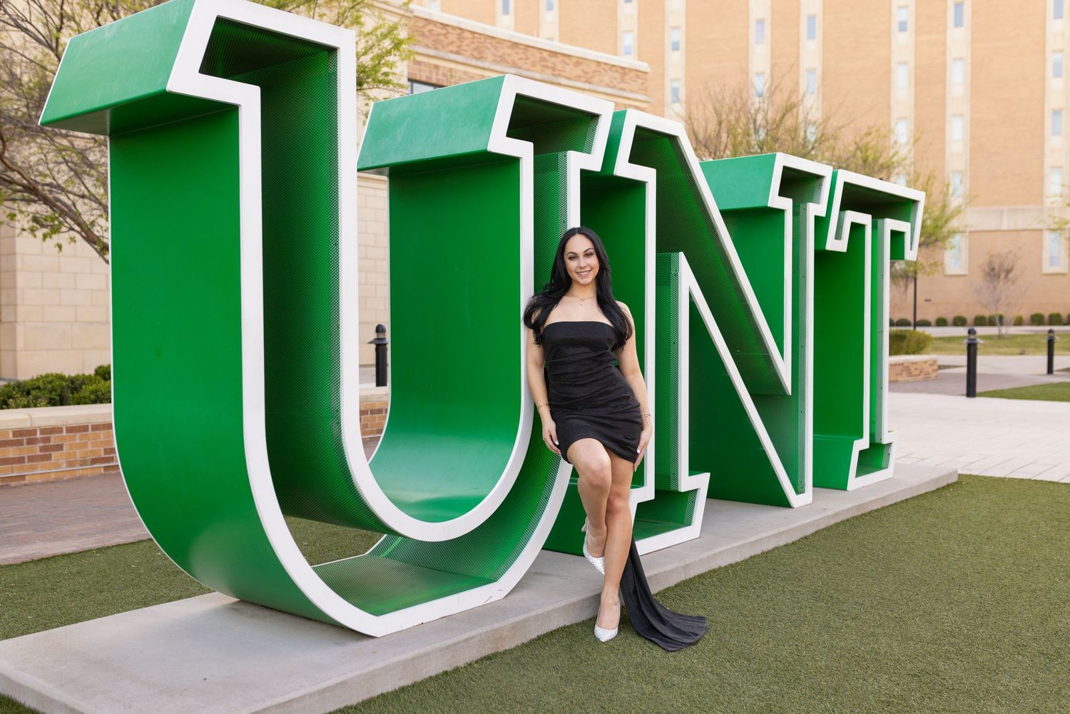 A person poses next to large green UNT letters outdoors on a university campus.