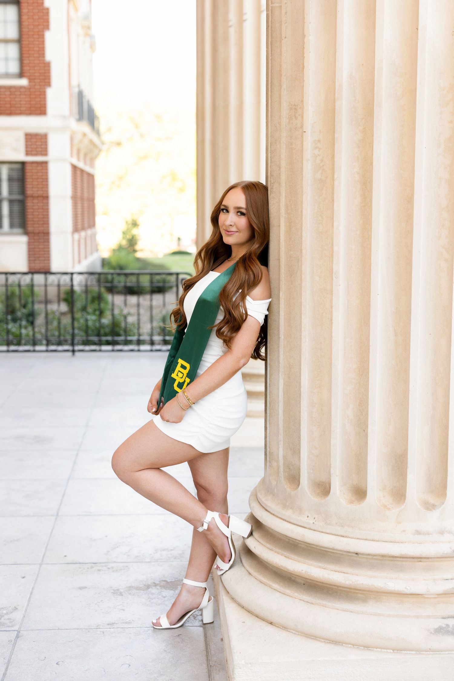 Graduate in white dress and green sash poses beside classical column on campus.