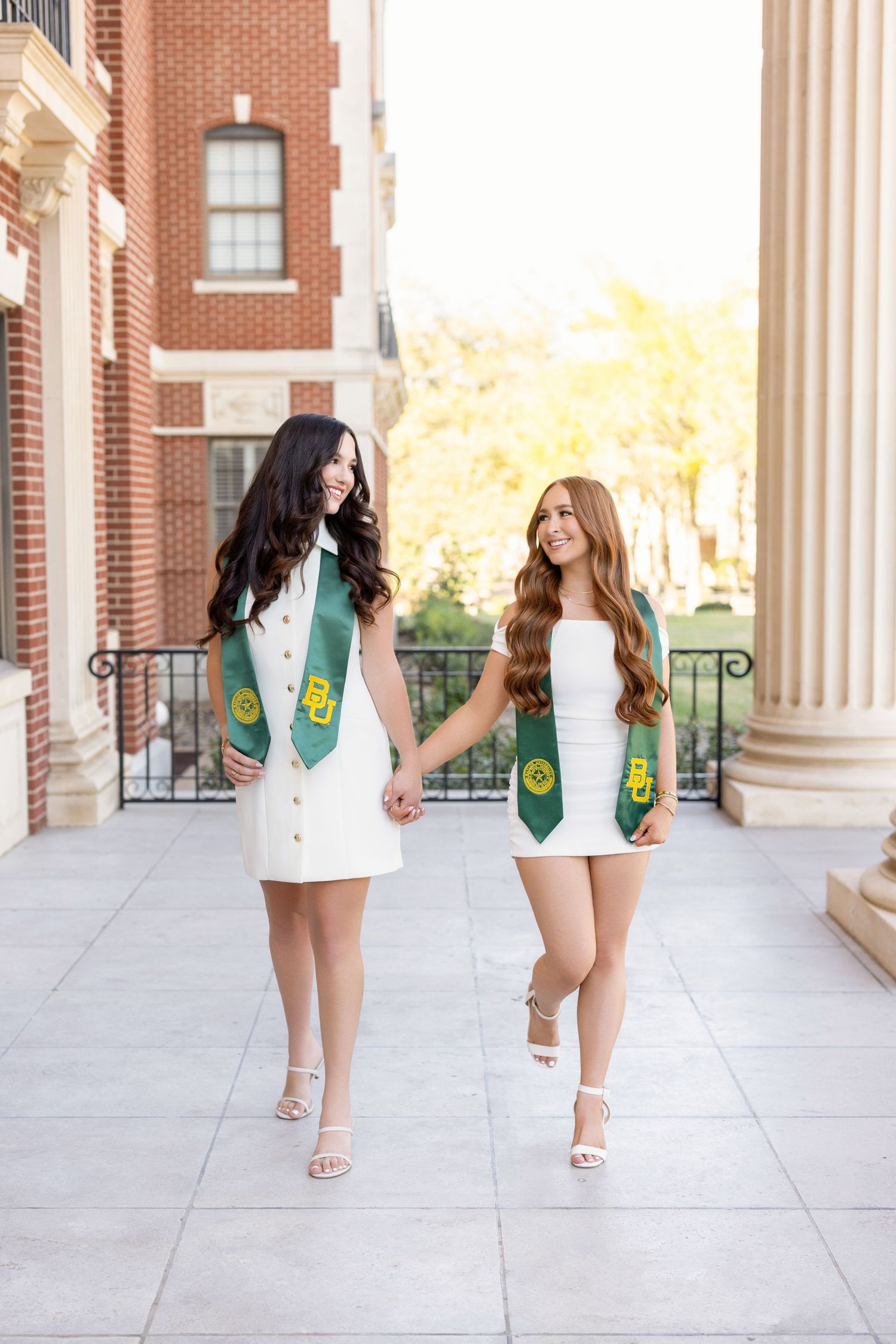 Two graduates in white dresses and green stoles with golden emblems walk together on a university campus sidewalk.
