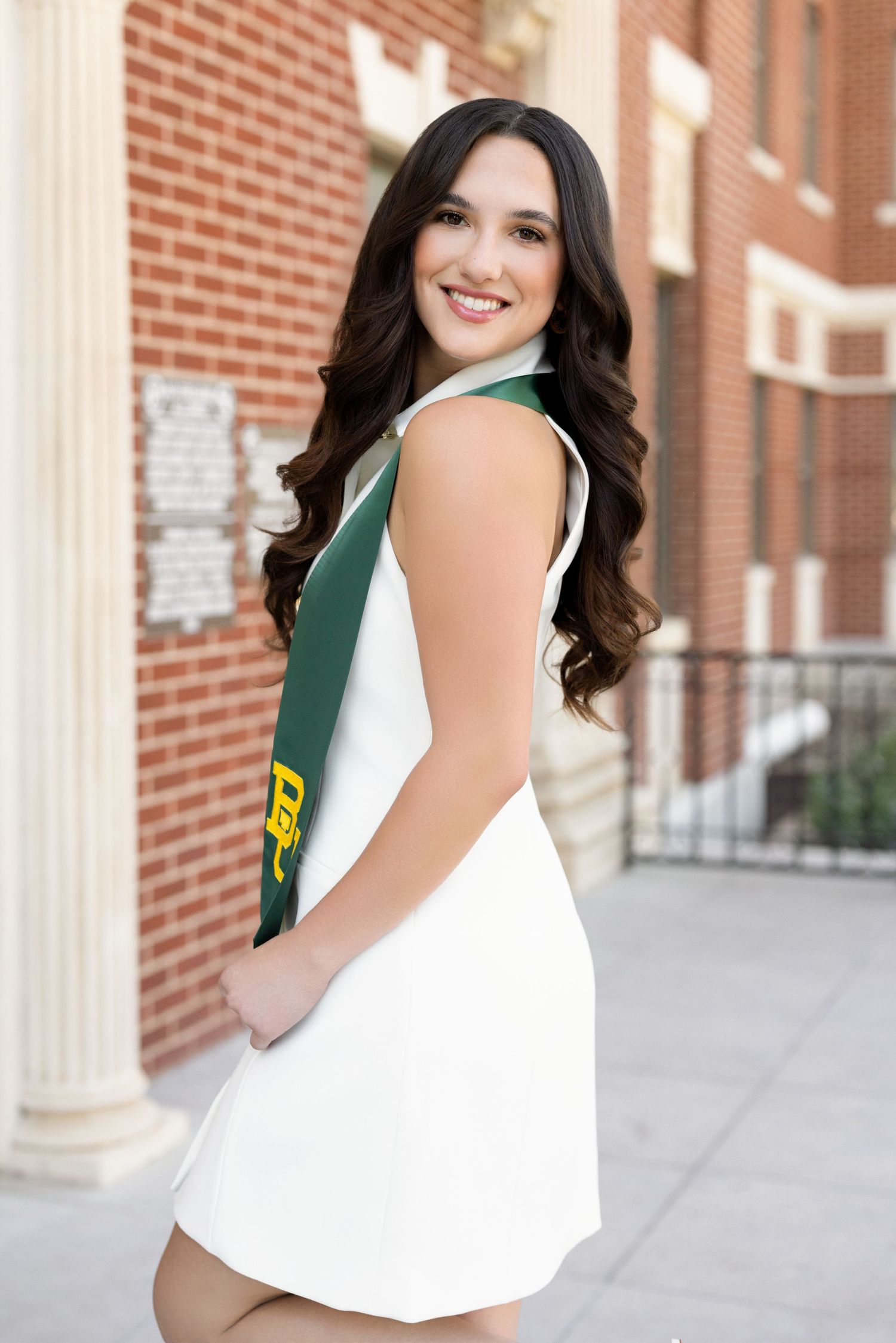 A graduate in a white and green dress poses against a brick wall during a senior photo session.