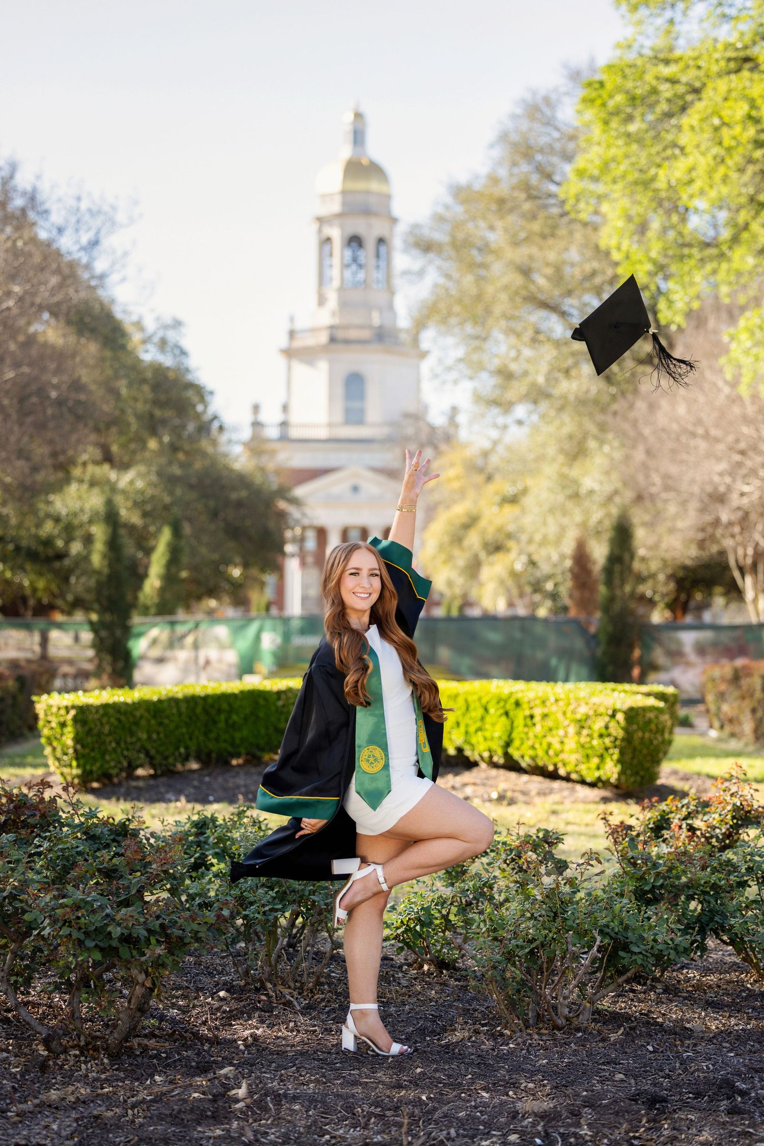 Graduate celebrating on campus with iconic chapel building and manicured gardens in background.