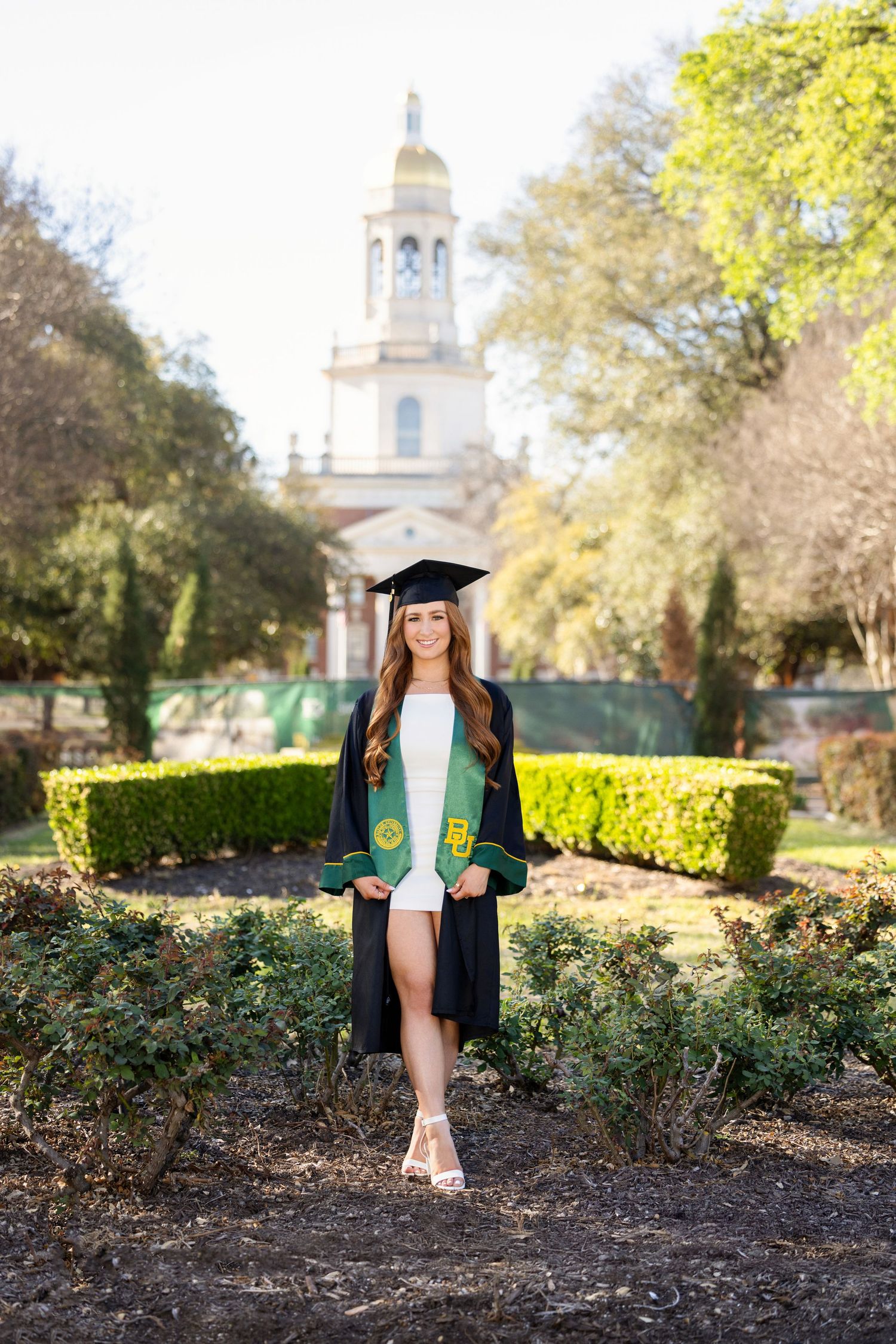 Graduate in cap and gown stands in front of grand university building with white spire and manicured gardens.