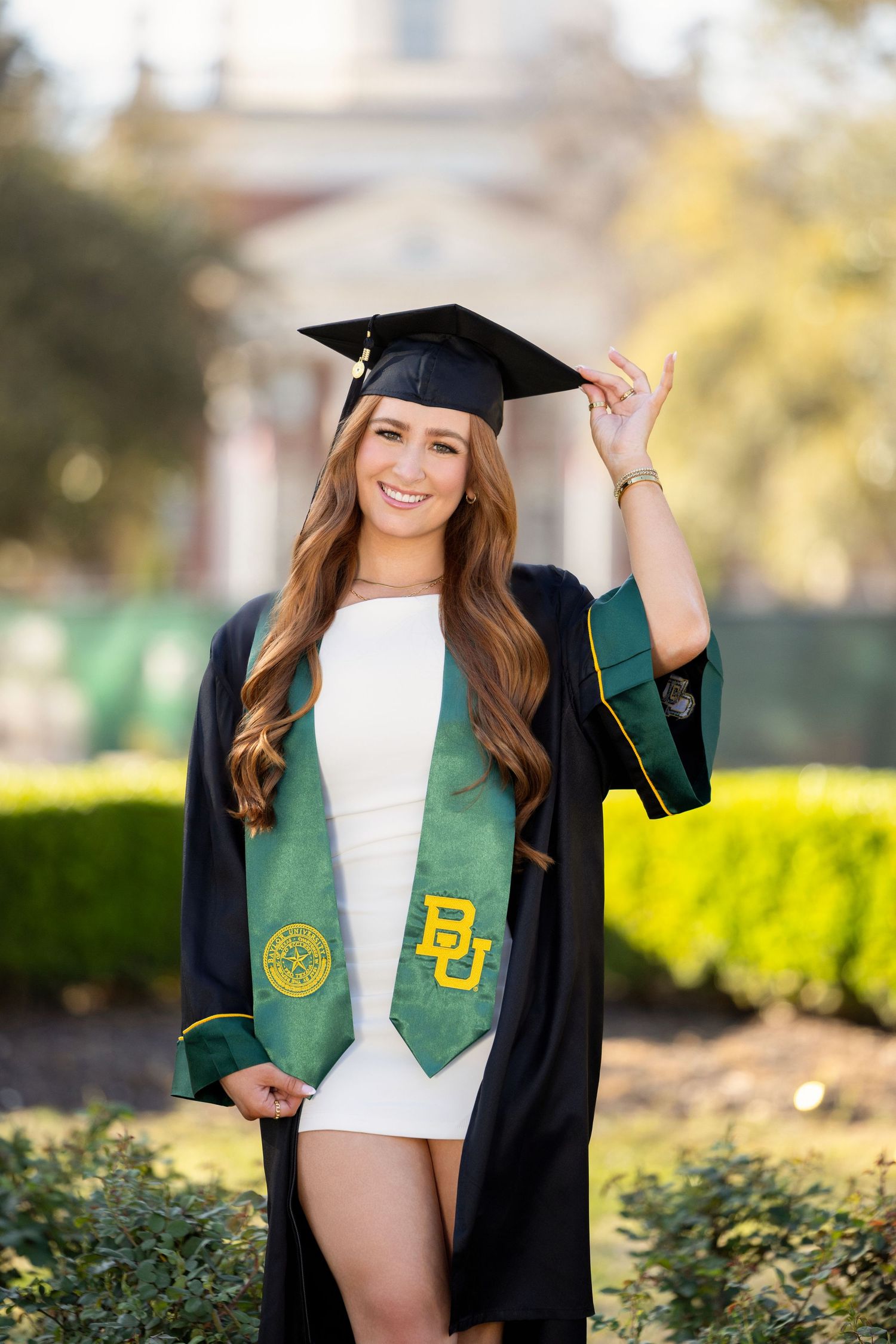 Graduate in cap and gown wearing green and black academic regalia from Baylor University on sunny day with landscaping.