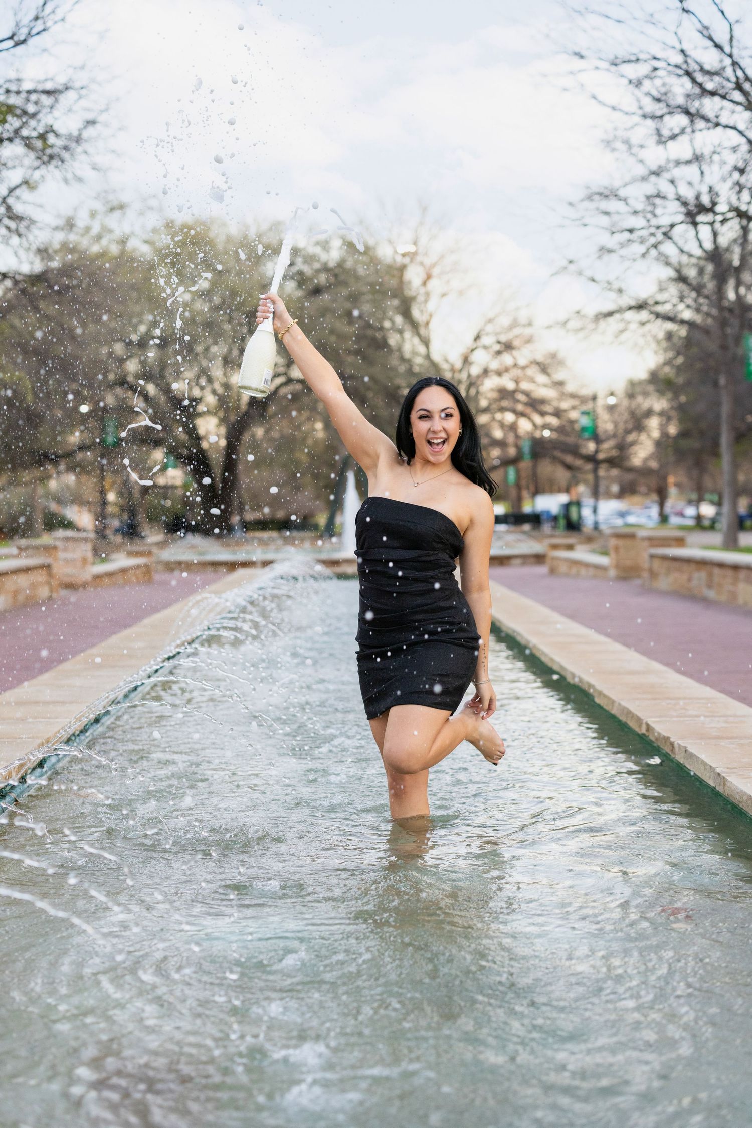Woman in black dress splashing and jumping joyfully in fountain water on a path during daytime.