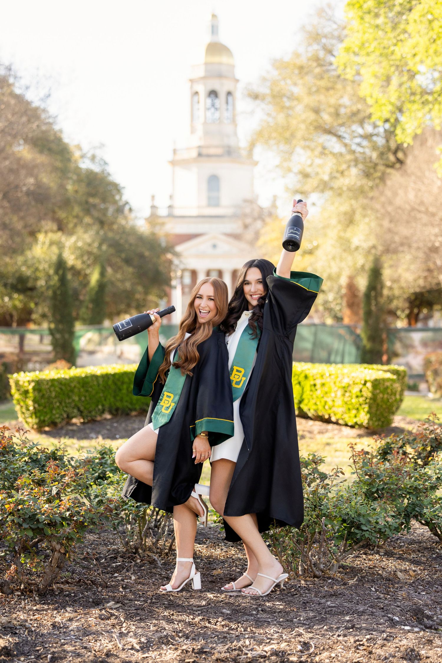 Two graduates in black gowns and green stoles celebrating together in front of historic university building.