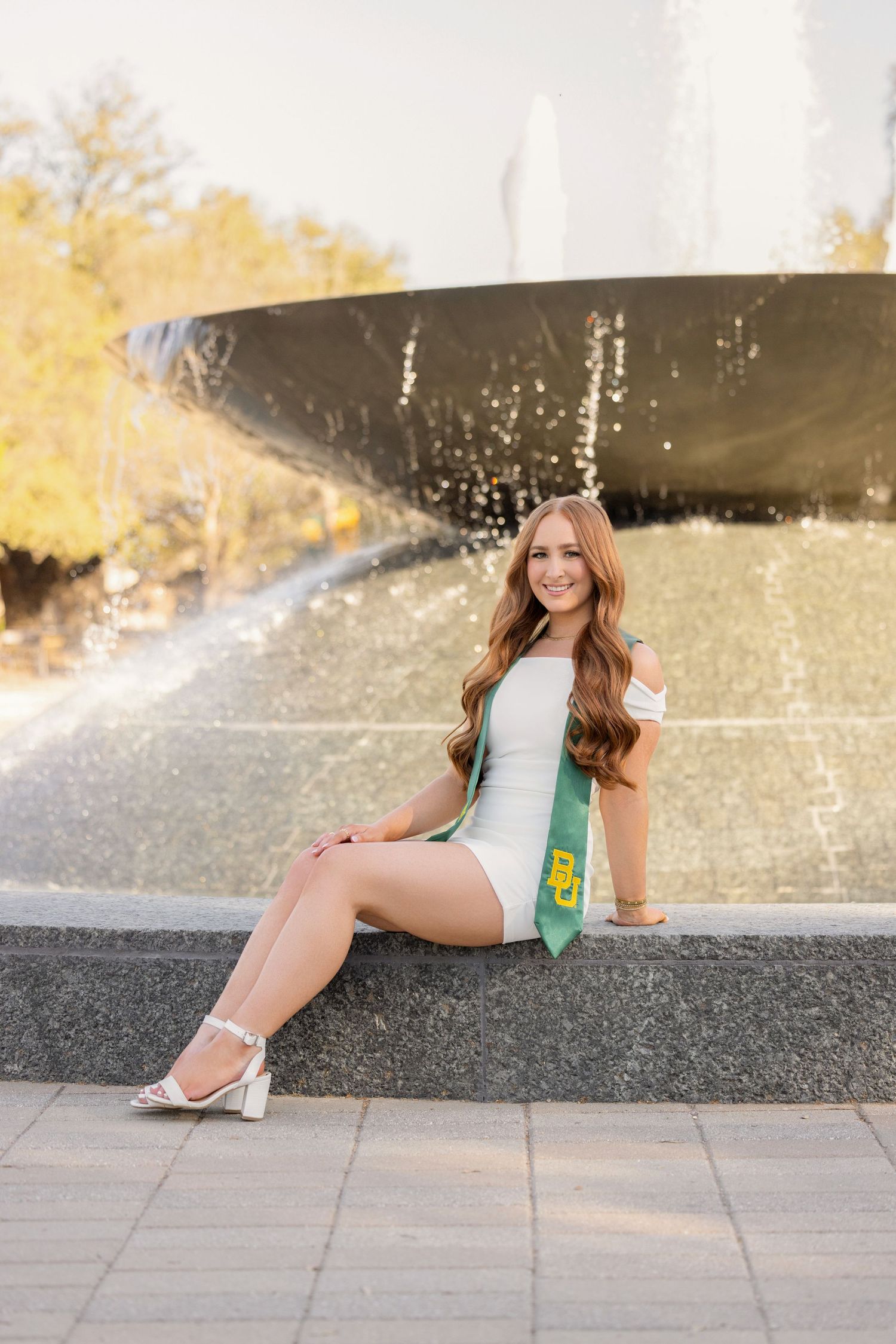 Graduate in white dress and green sash poses by fountain during sunny graduation photoshoot.