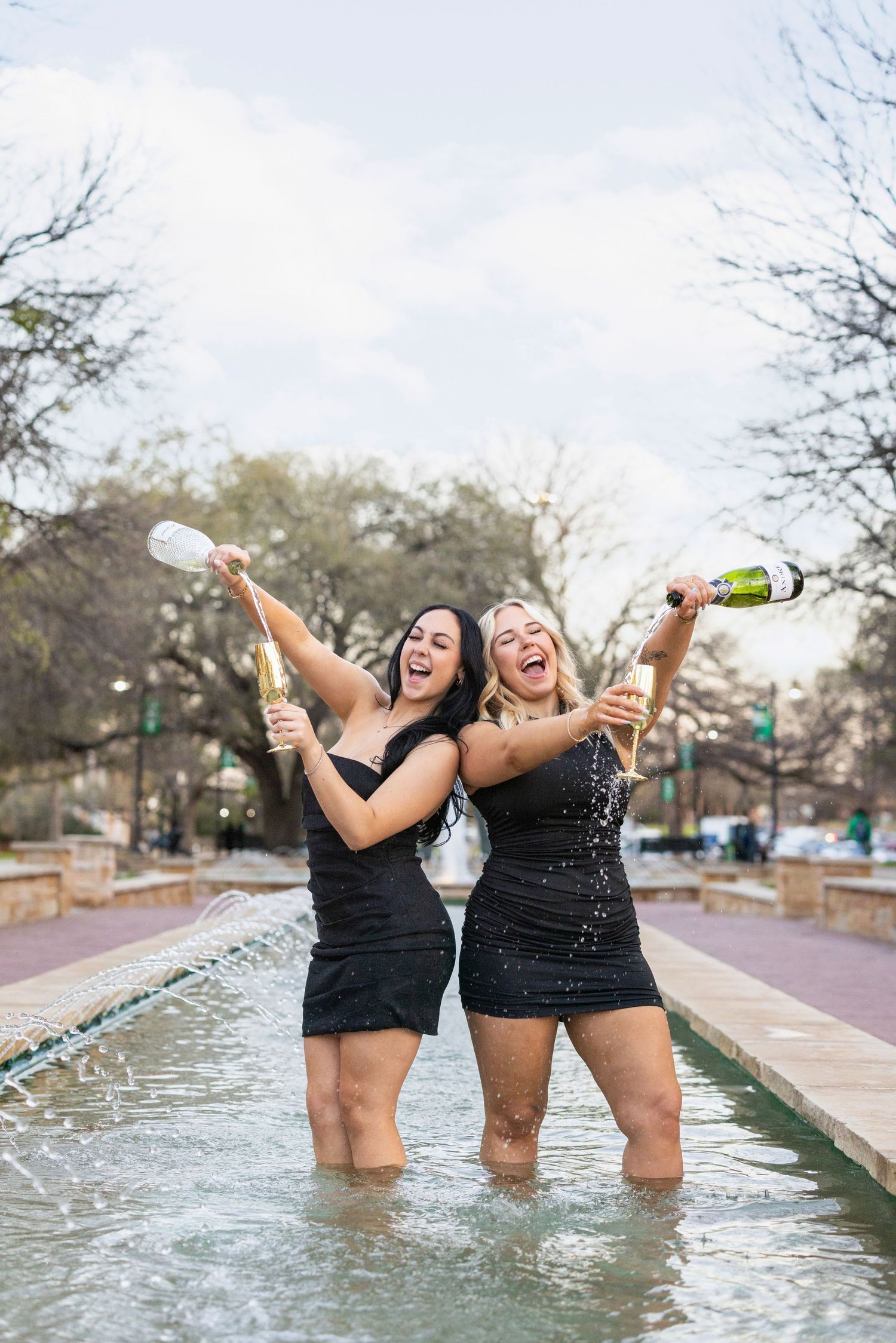 Two people in black dresses spray champagne while celebrating and standing in a shallow fountain.