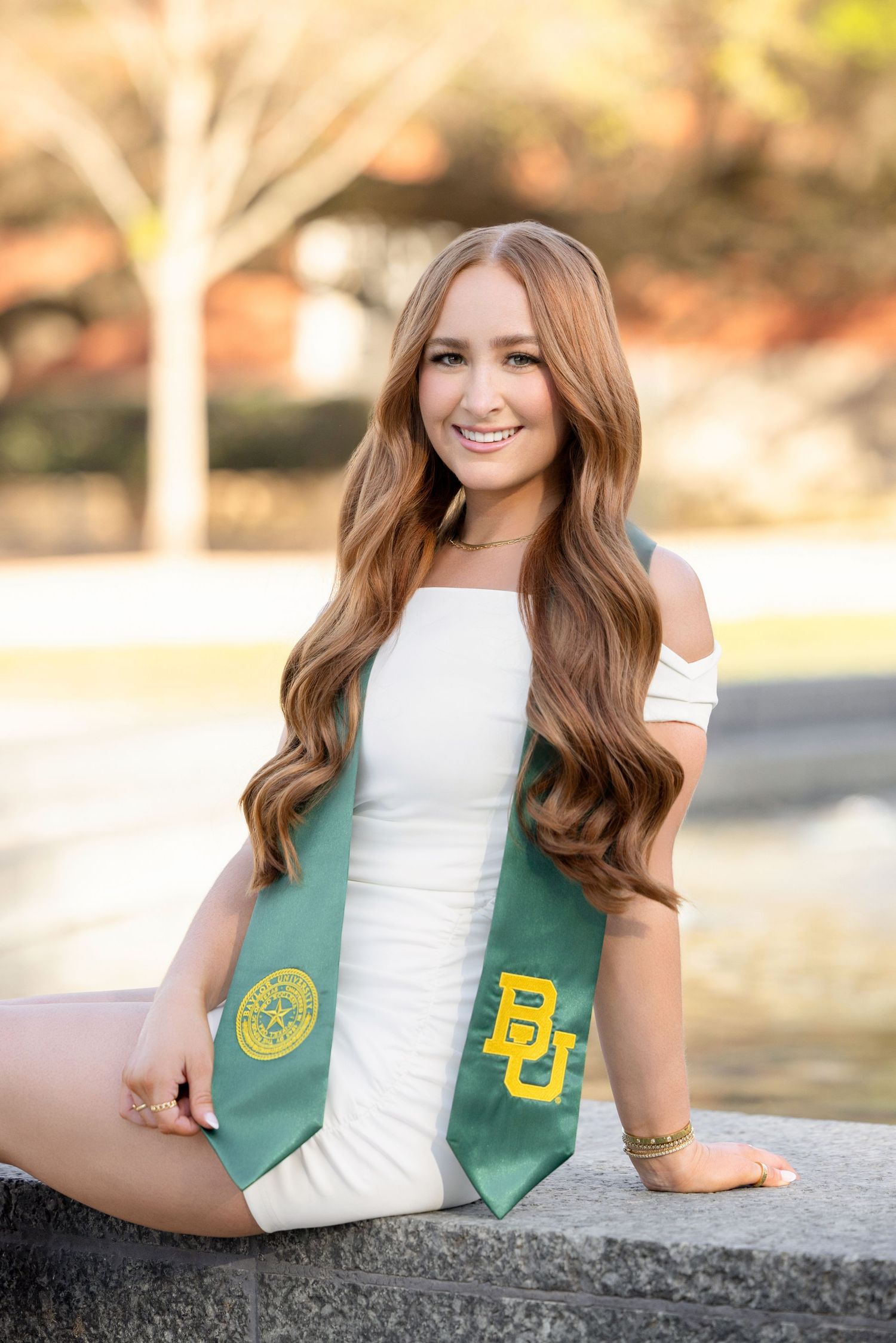 Graduate wearing white off-shoulder dress and green Baylor University graduation stole poses outdoors on sunny day.