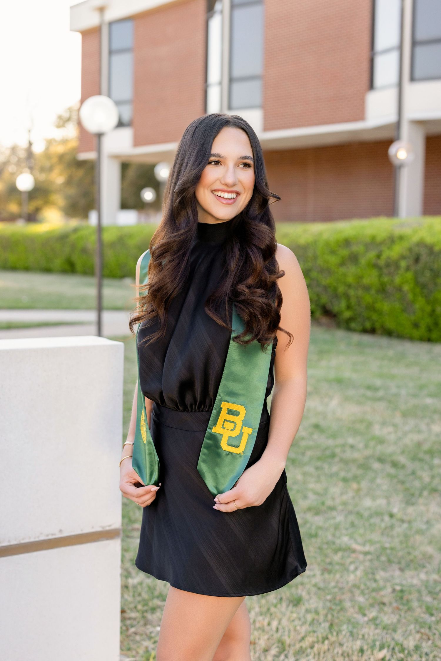 Student wearing a black dress and green Baylor University graduation stole poses outdoors on campus during sunset.