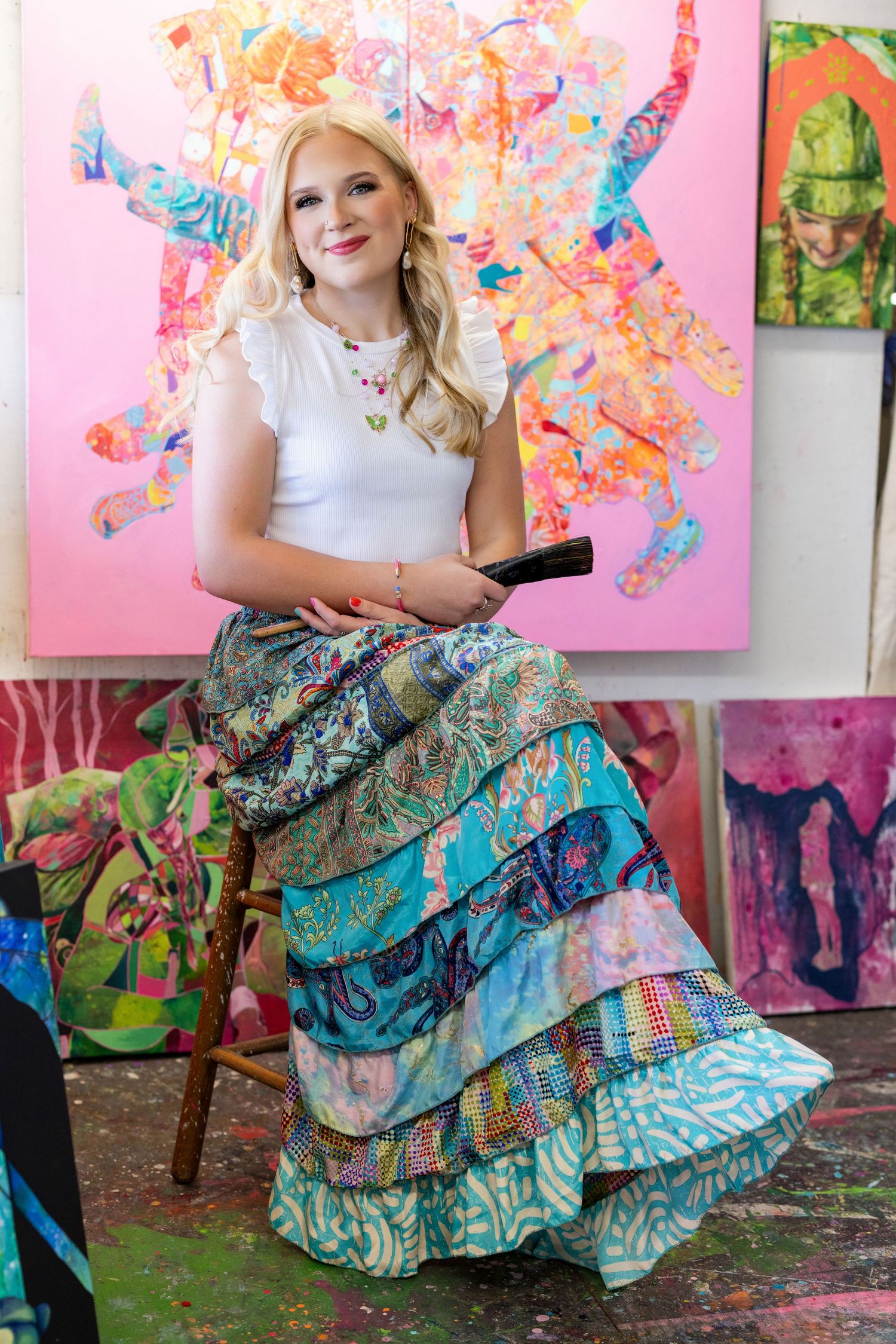 Artist sits in colorful studio with vibrant tiered maxi skirt against backdrop of abstract paintings.