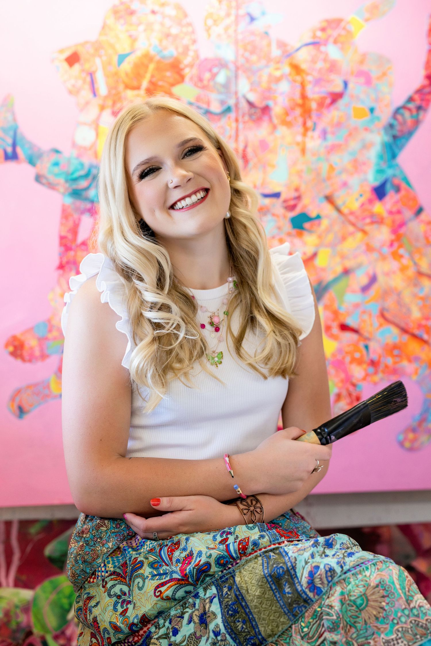 A creative artist in a white top and patterned skirt smiles while holding art supplies against vibrant artwork.