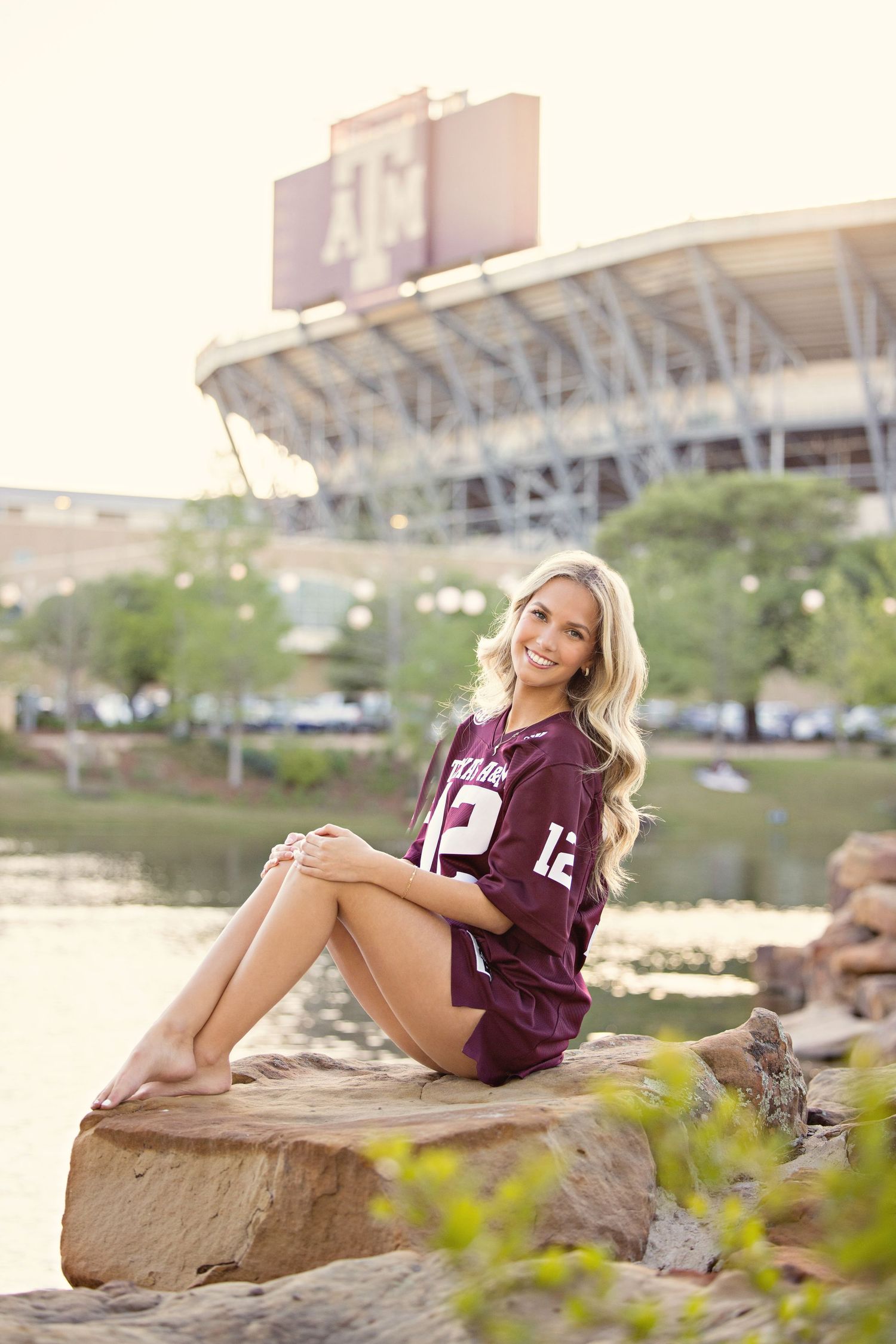 College sports fan in maroon jersey poses on rock near stadium during golden hour outdoor photo session.