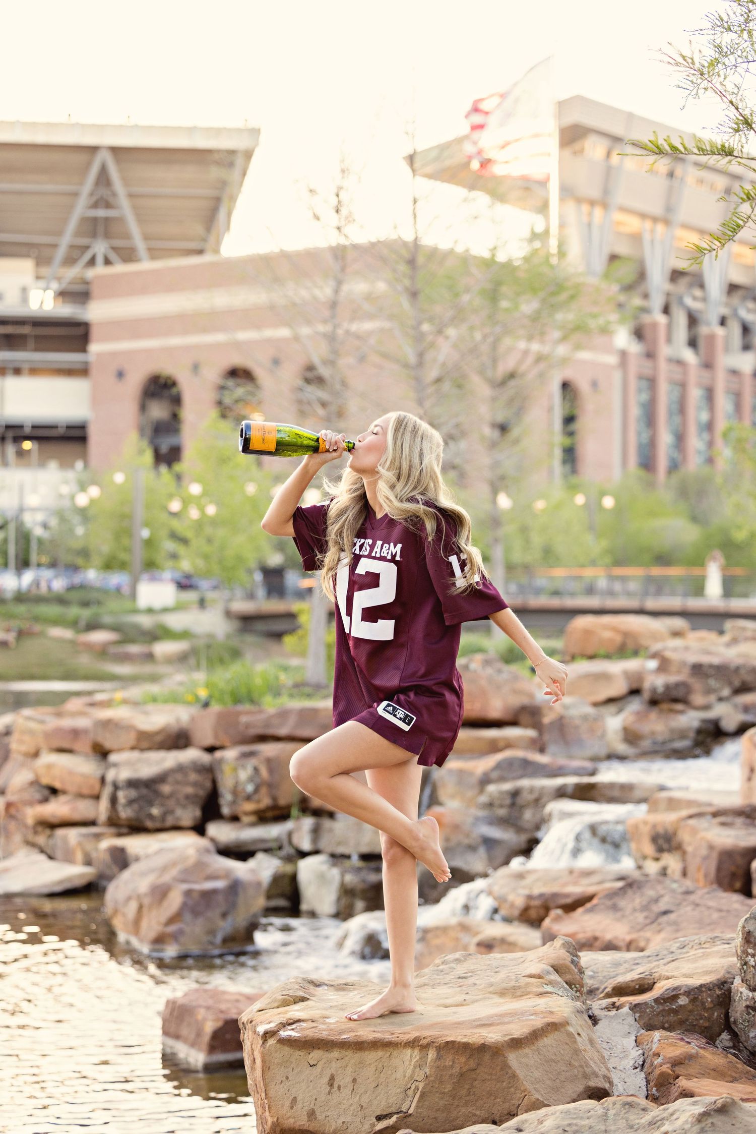 A person in a maroon Texas A&M jersey stands on rocks by a water feature drinking from a bottle.