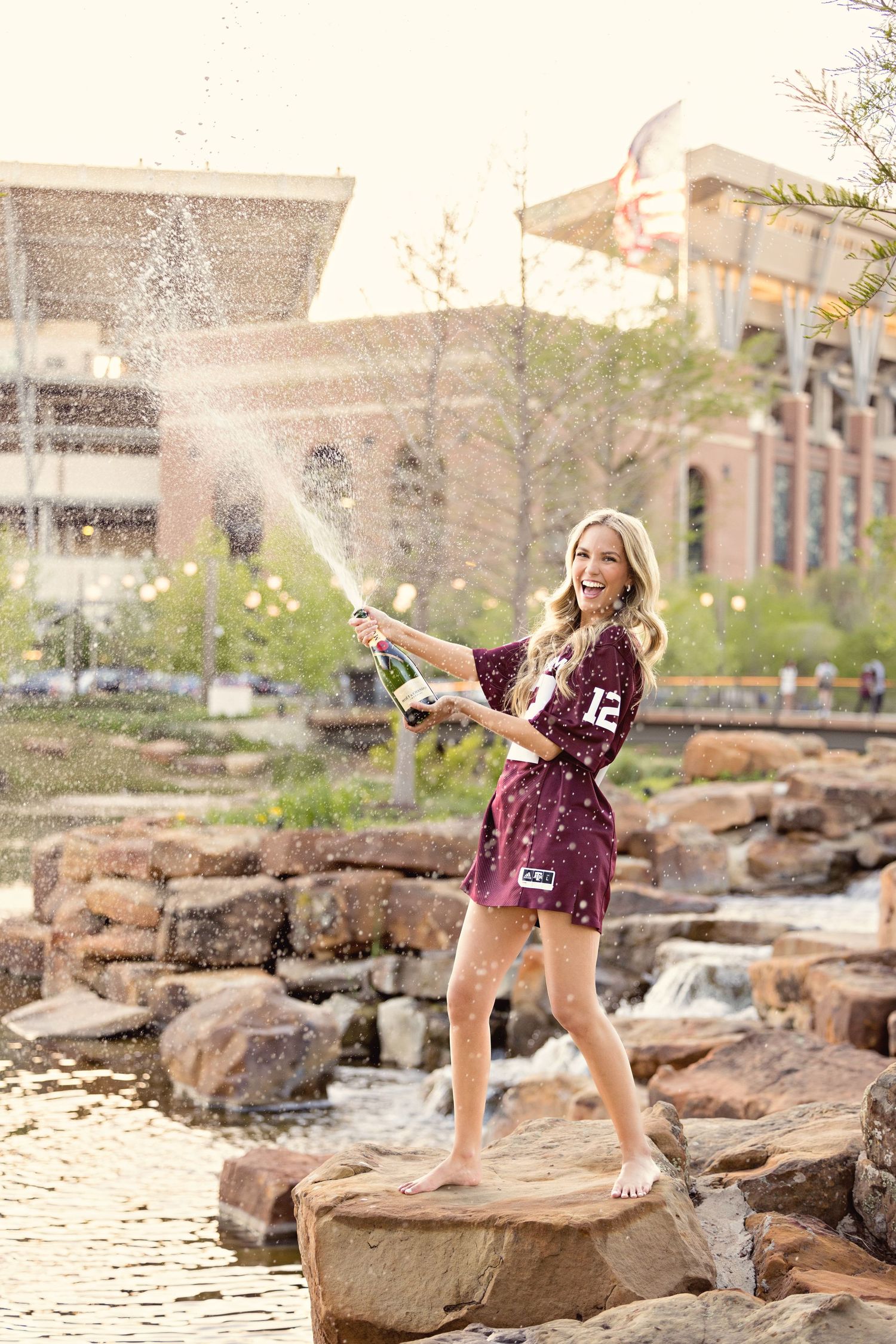 Student in maroon jersey sprays champagne while standing on rocks by a water feature on a sunny day.