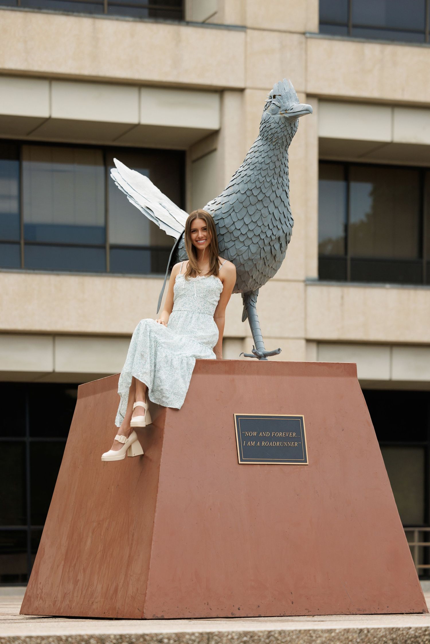 Person in a white dress sits casually on a large brown pedestal beside a metallic bird sculpture outside a building.