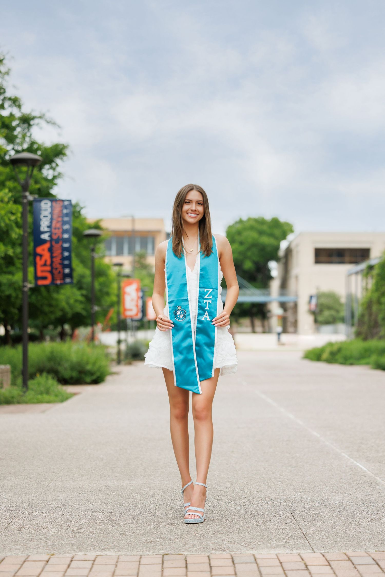 A young graduate wearing a turquoise and white stole stands on a university campus pathway with a bright smile.