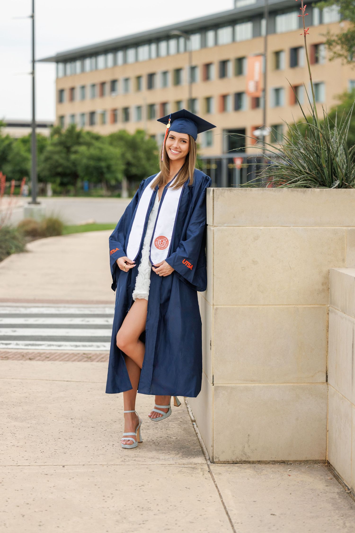 Graduate in navy blue cap and gown poses outside a brick university building on a sunny day.