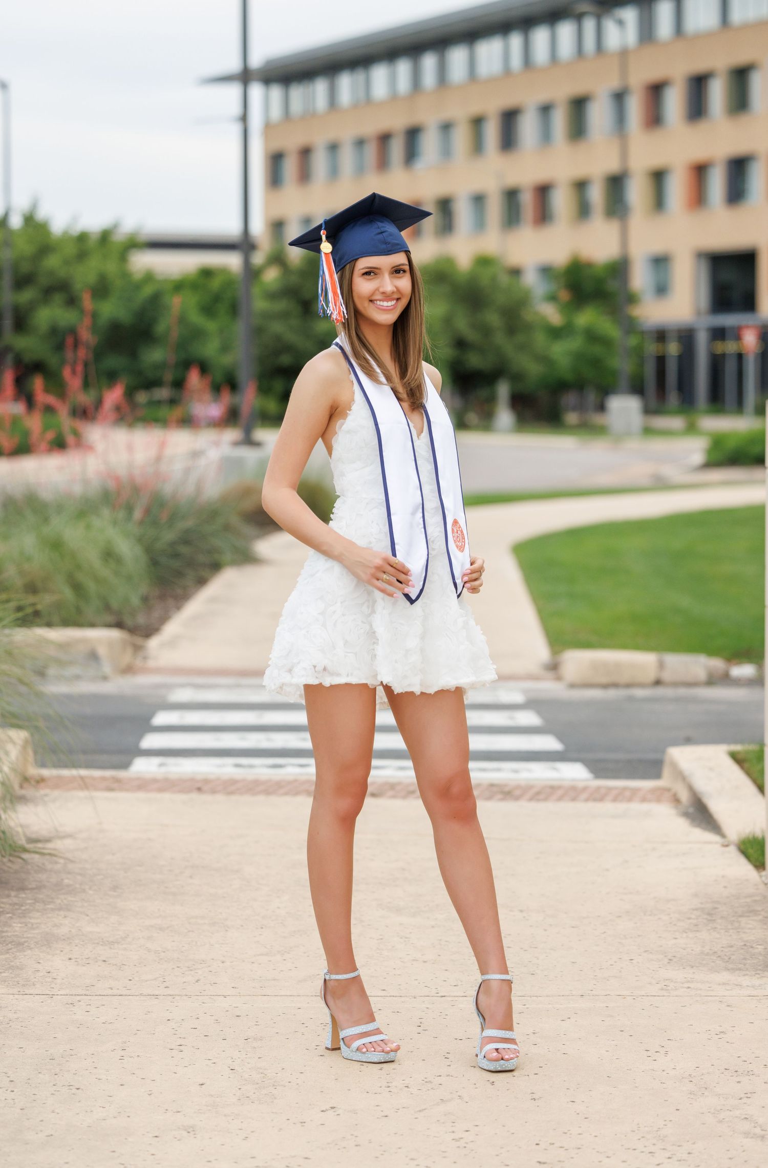 Graduate in blue cap and white dress poses on campus walkway with building and landscaping in background.