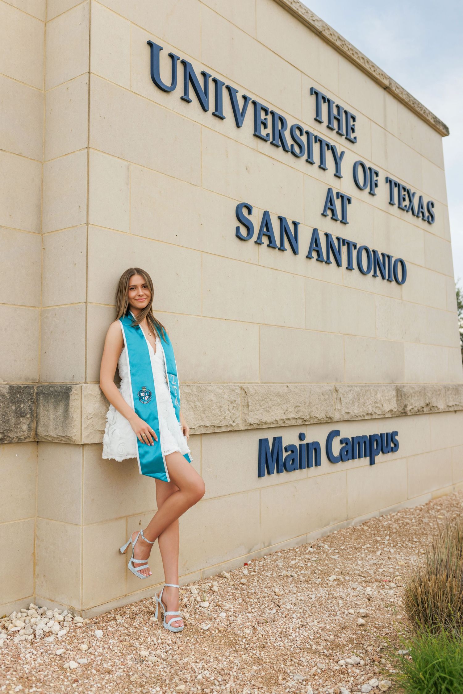Graduate poses in front of The University of Texas at San Antonio Main Campus entrance sign wearing graduation stole.