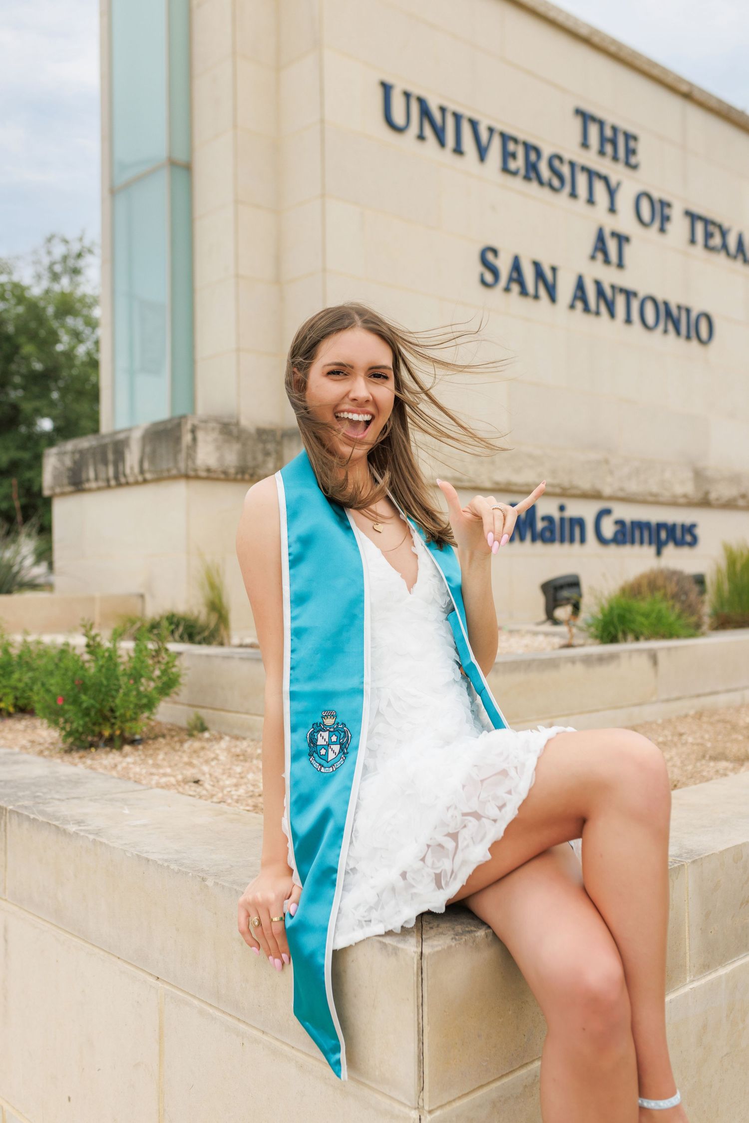 Graduate in white dress and blue graduation stole sits on steps at The University of Texas at San Antonio main campus.