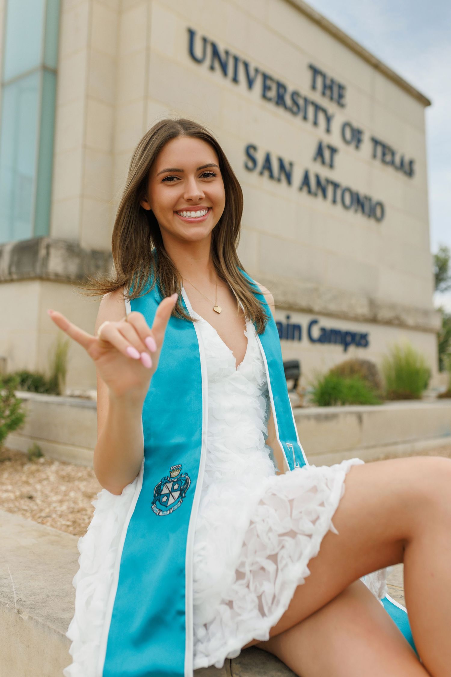 A graduate in a turquoise graduation stole and white dress poses with a peace sign at the University of Texas at San Antonio.