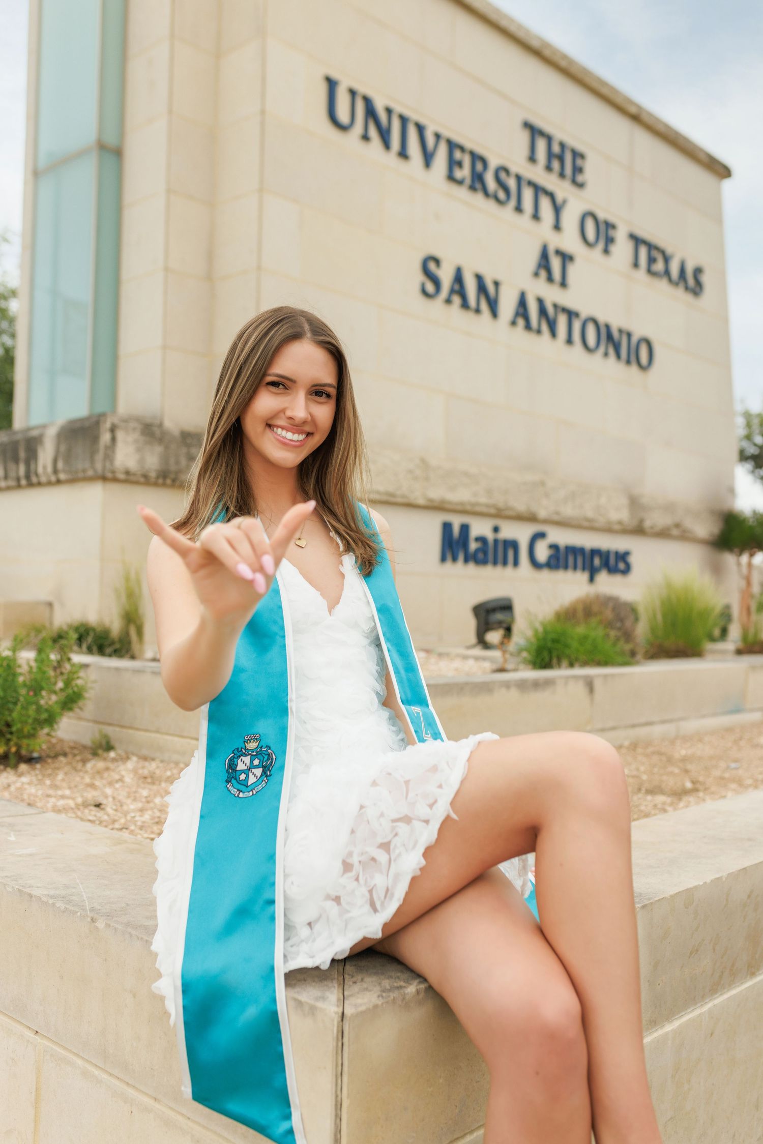 Graduate celebrates at the University of Texas at San Antonio Main Campus entrance wearing graduation stole and white dress.