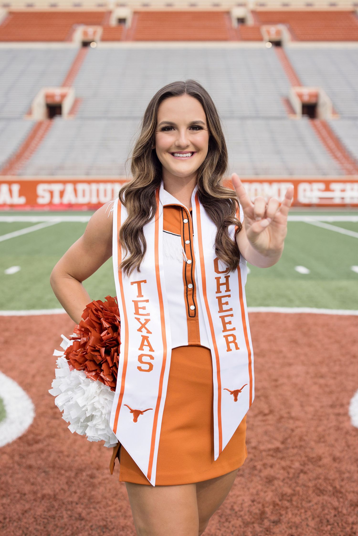 University of Texas cheerleader wearing orange uniform and graduation sash poses with pom poms at football stadium.
