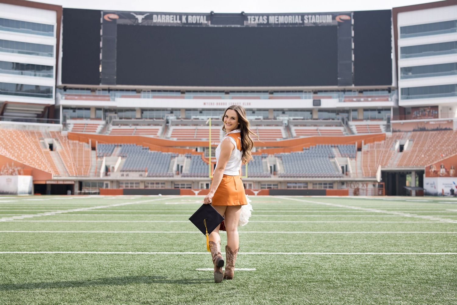 A cheerleader performs on the field at a large stadium with empty orange bleachers and scoreboard in the background.