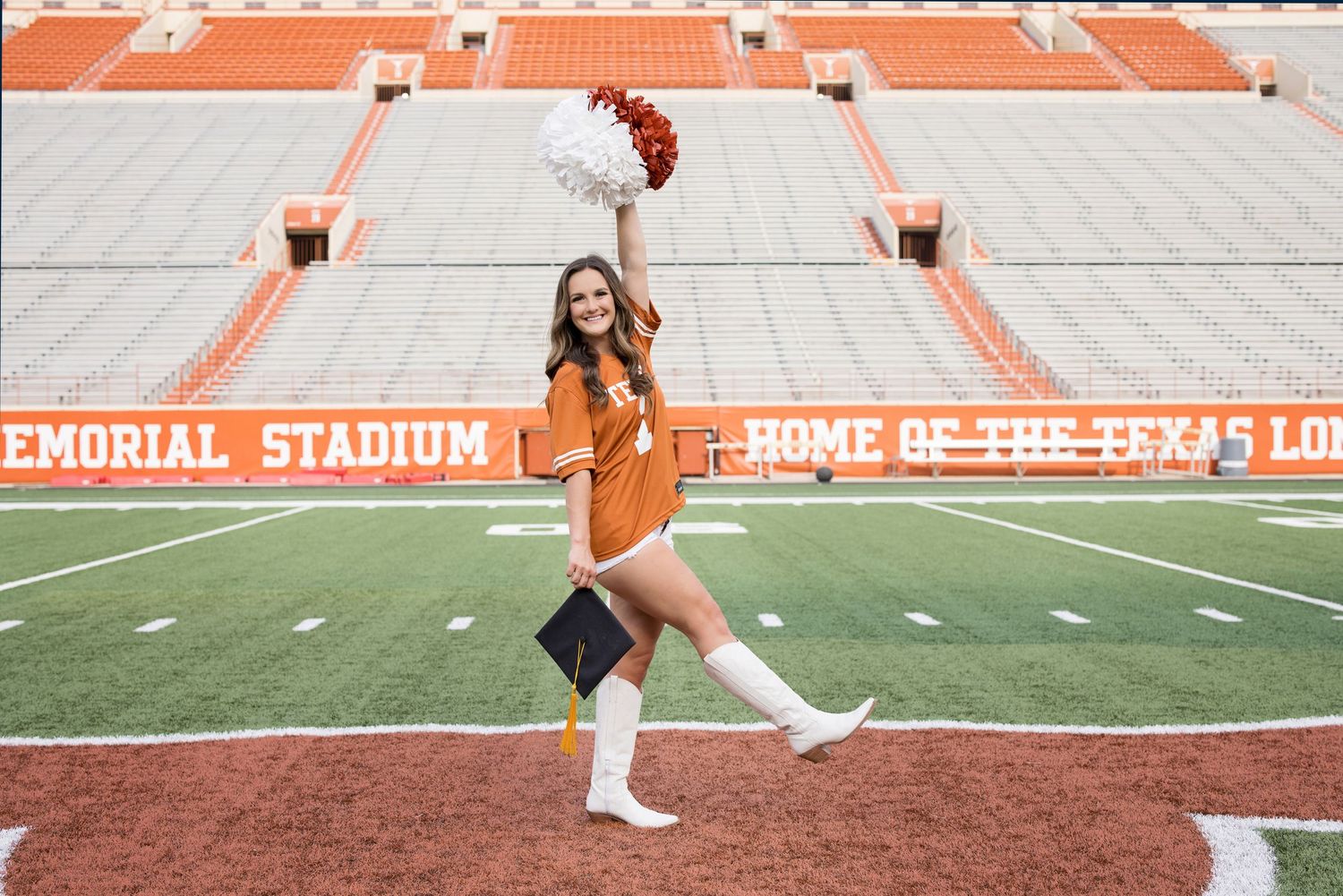 A graduate in a burnt orange shirt and white boots celebrates with pom poms at Texas Memorial Stadium.