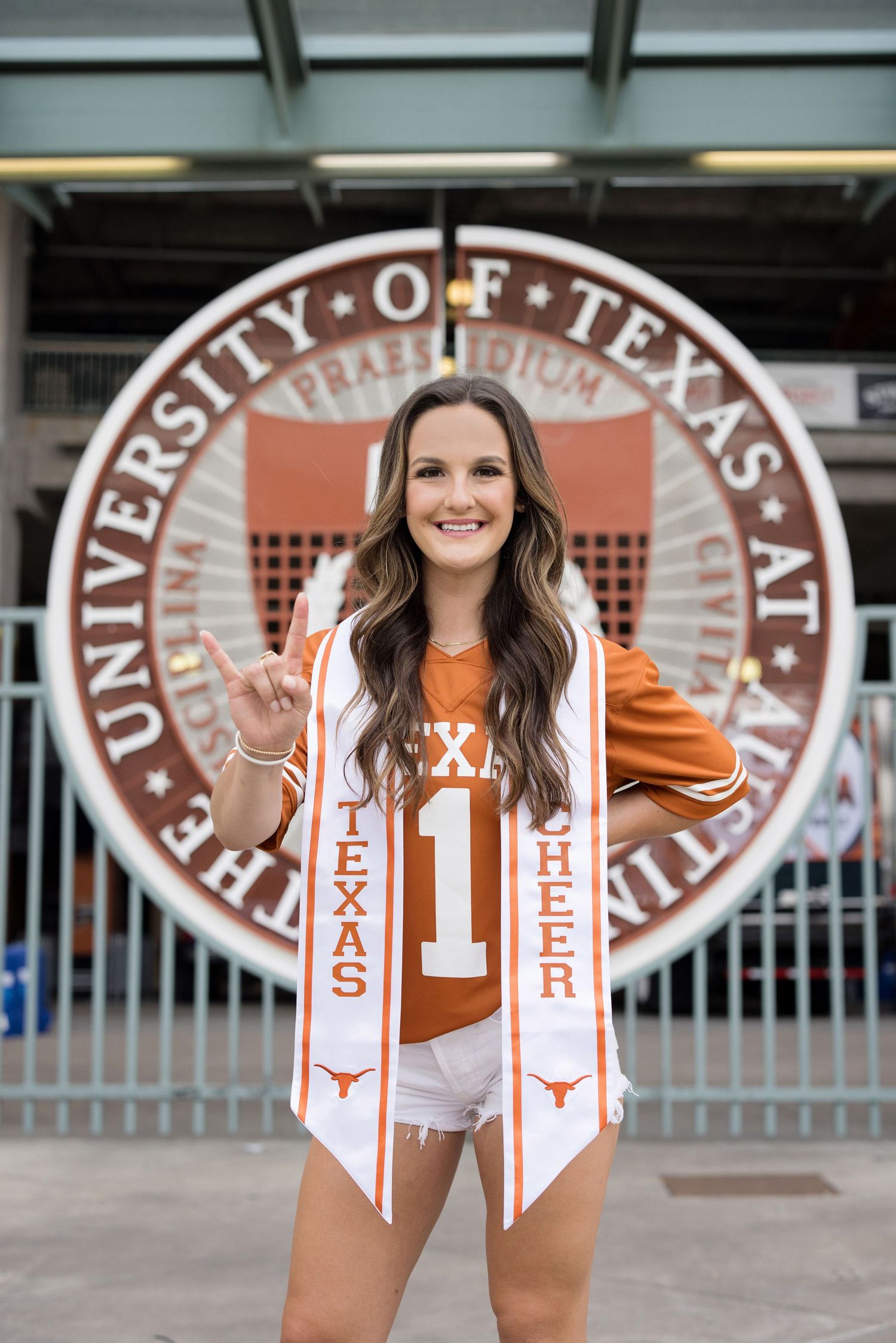 A University of Texas graduate poses in front of the UT seal wearing a burnt orange jersey and graduation stoles.