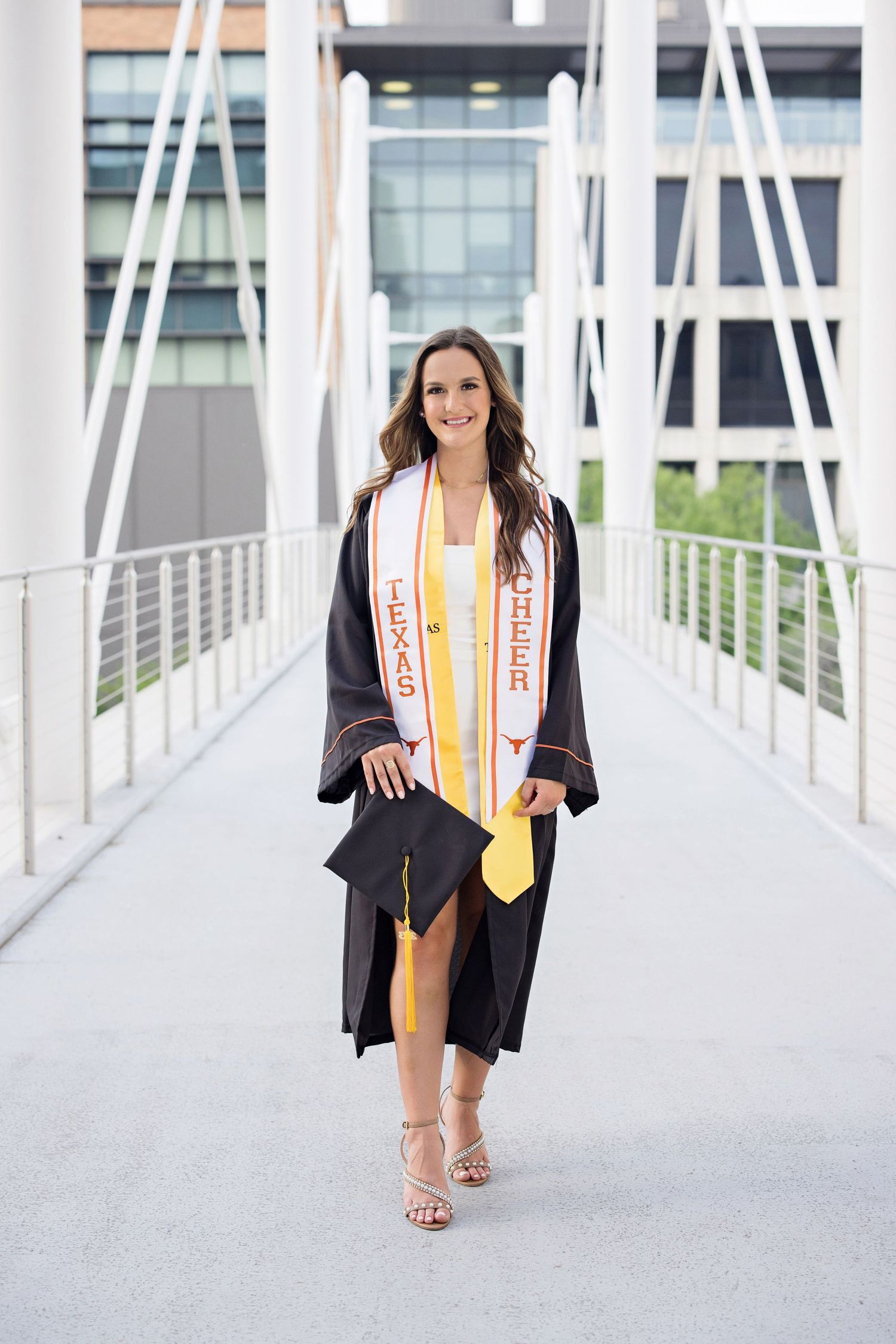 Graduate in black cap and gown with honors sash walks confidently on modern white bridge walkway.