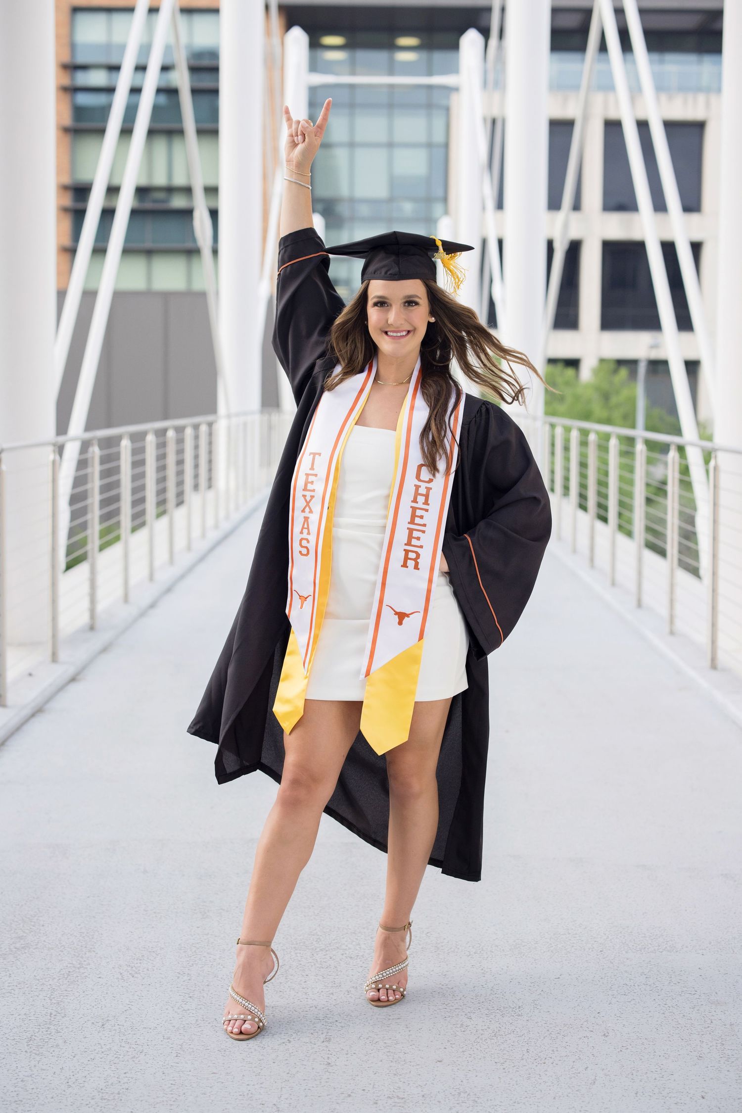 A graduate in cap and gown poses on a modern bridge walkway with a peace sign gesture and graduation stole.