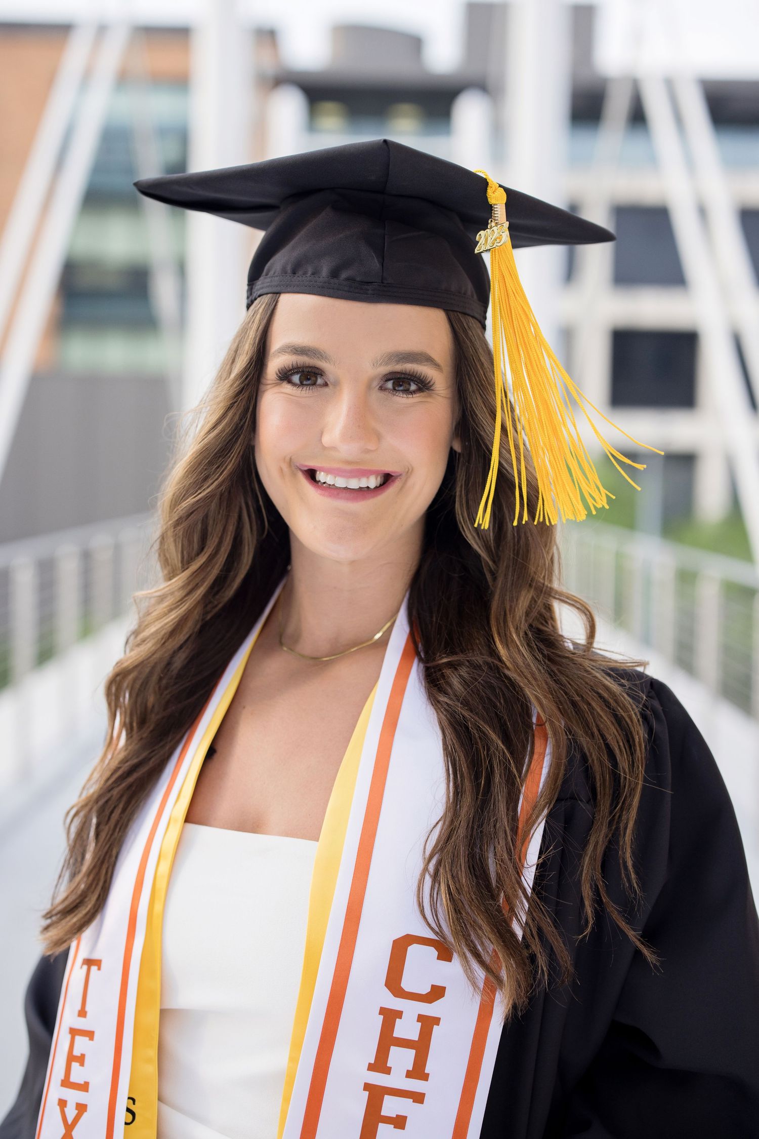 Graduate in cap and gown wearing a white dress and ceremonial sash with yellow tassel smiles in graduation photo.