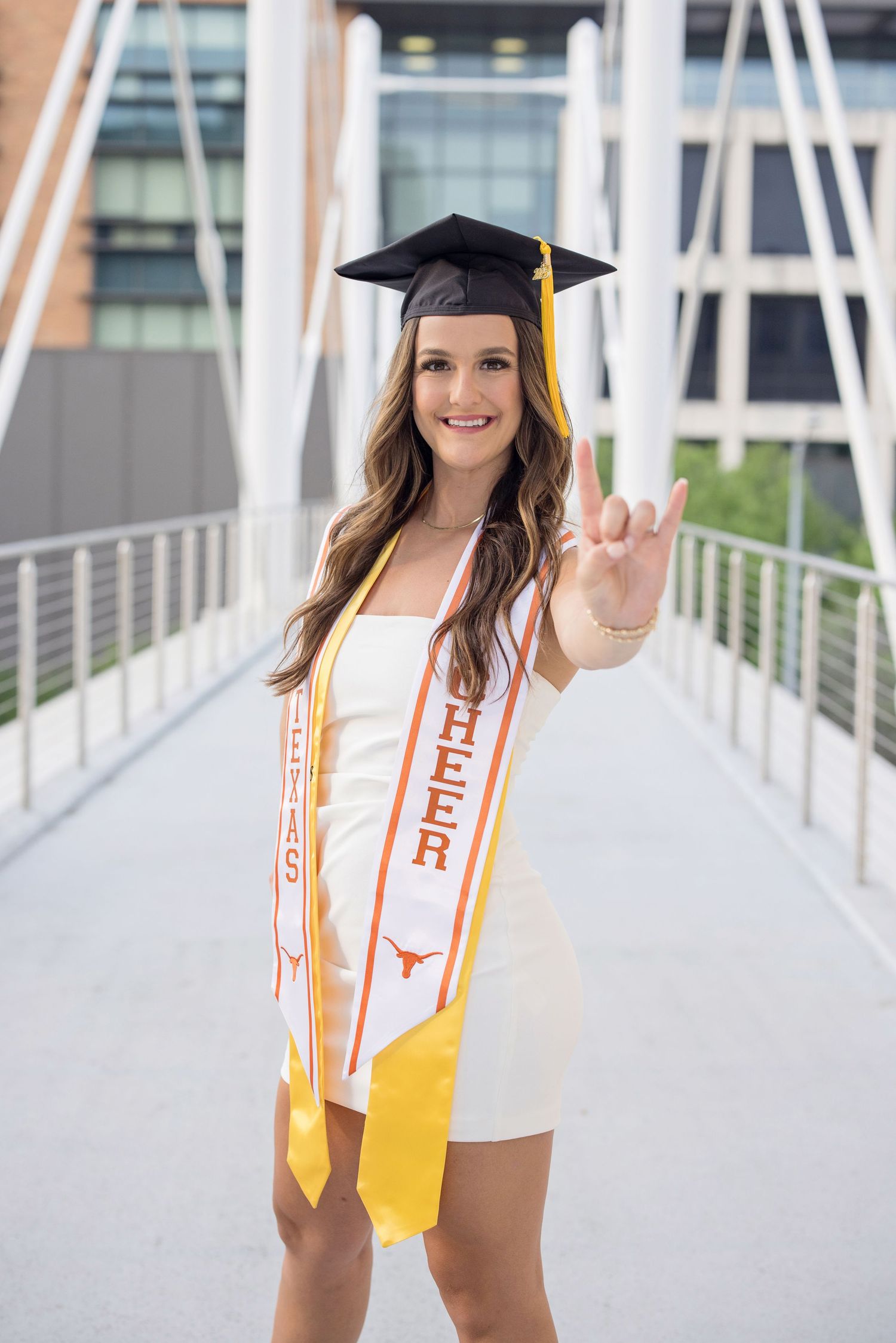 Graduate in black cap and white dress with orange University of Texas sash poses on modern bridge.