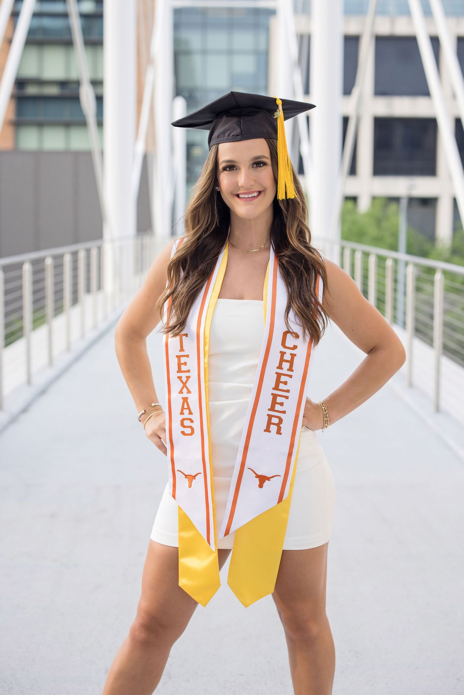 Graduate in white dress and cap with University of Texas graduation sashes poses on modern walkway bridge.