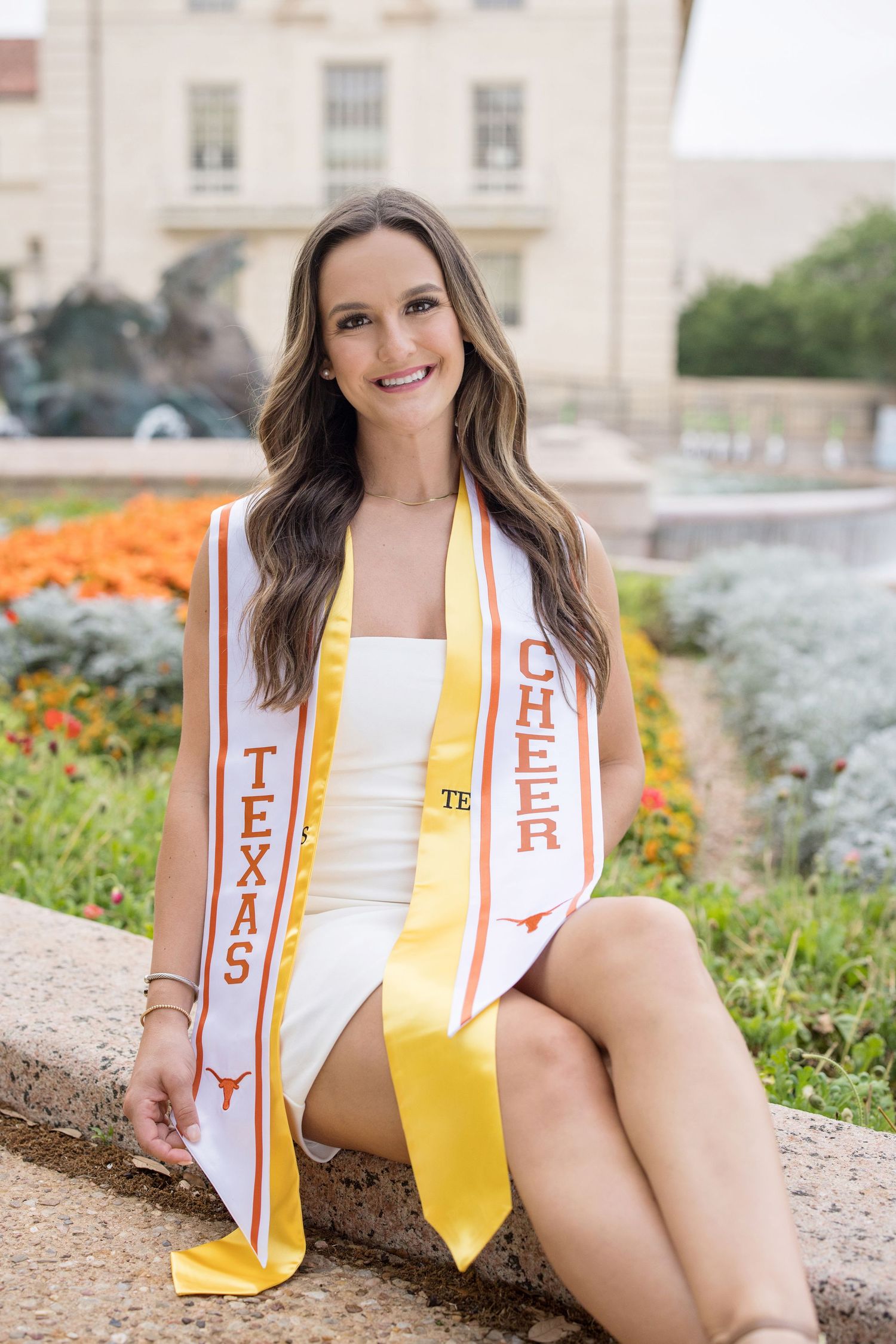 Graduate in white dress and orange-and-white stole poses on campus with flowers in background near fountain.