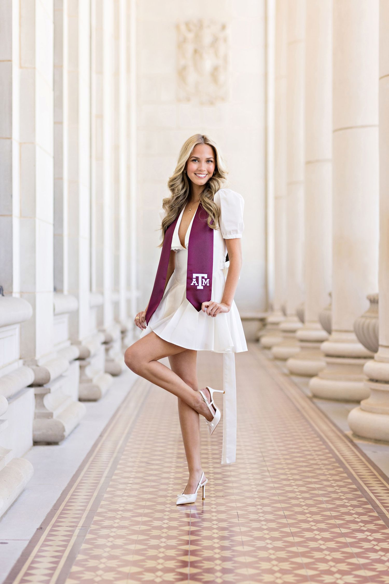 Graduate in white dress and maroon Texas A&M stole poses between classical columns with tiled floor.