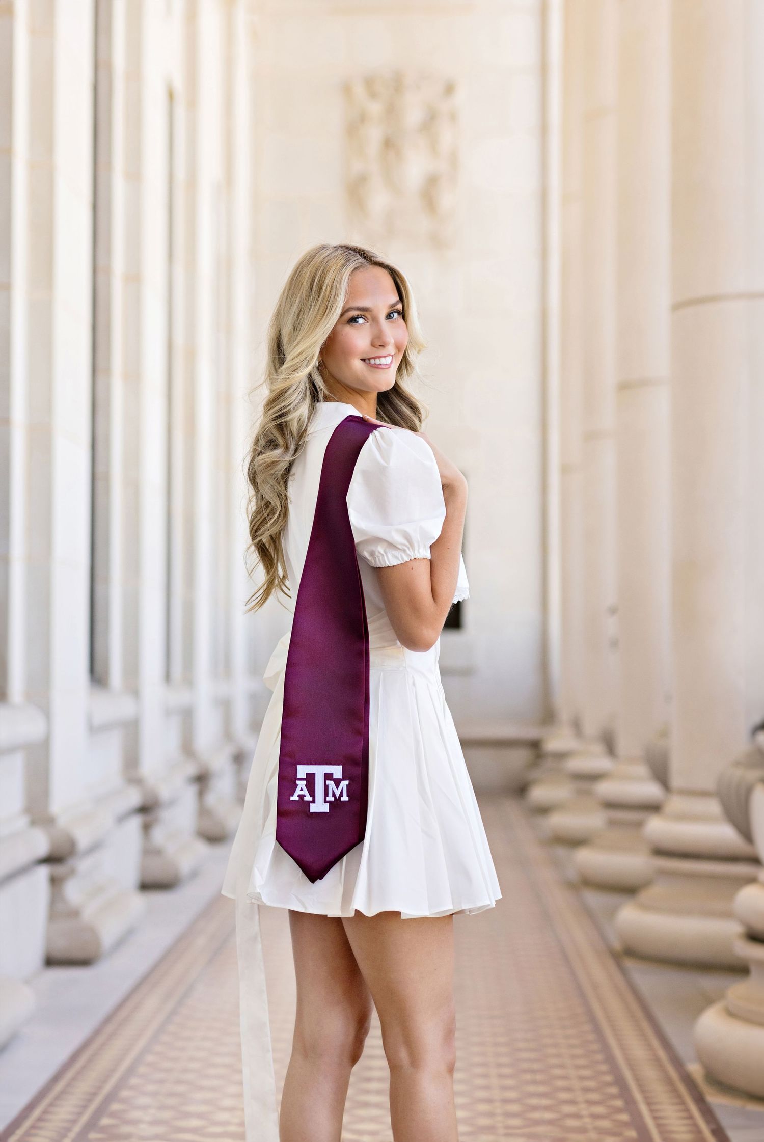 A person wearing a white dress and maroon Texas A&M University necktie poses in an elegant columned hallway.
